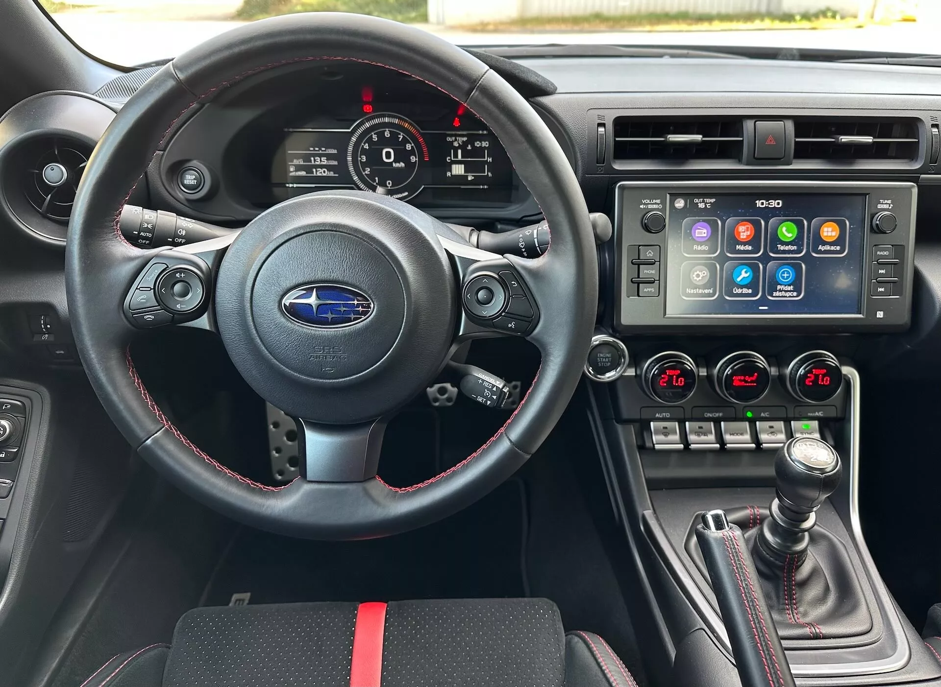 Interior do Subaru BRZ 2023 pela perspectiva do motorista incluindo um volante desportivo em pele com controles multifunções e costura vermelha, um cluster de instrumentos com mostradores analógicos e um display digital, uma consola central com um amplo sistema de infotainment touchscreen e controle climático de duas zonas, e uma alavanca de mudanças manual com capa de pele e acentos vermelhos, contra um interior preto com accentos desportivos vermelhos, realçando as características avançadas do veículo e tecnologia.