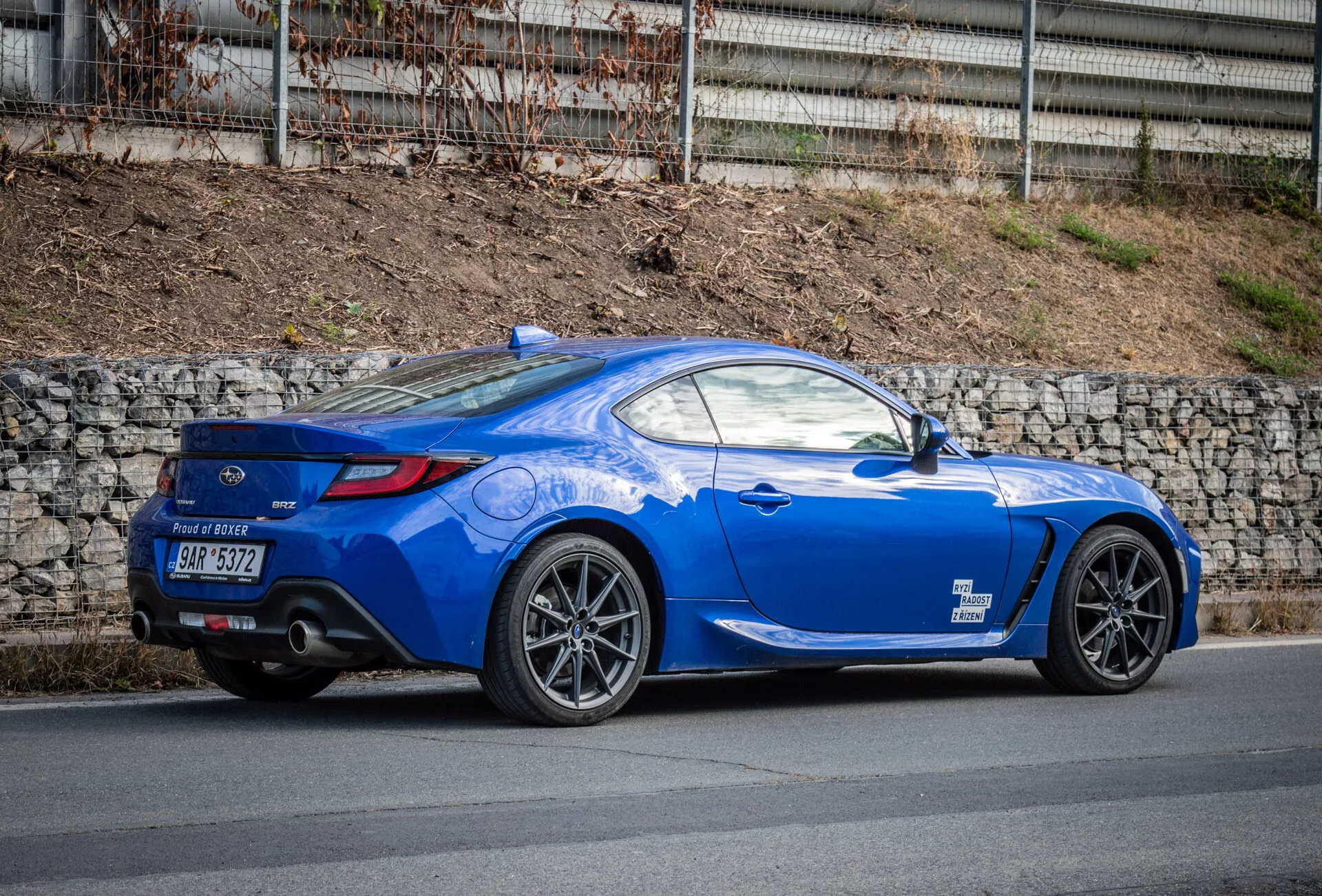 Coupé esportivo Subaru BRZ 2023 de cor azul brilhante com design aerodinâmico estacionado em uma rua, destacando o arco da roda traseira, lanternas traseiras, curvatura da janela traseira para a tampa do porta-malas, linhas esculpidas, linha do telhado inclinada, rodas de liga de múltiplos raios, freios de alto desempenho, spoiler traseiro de perfil baixo, tubos de escape duplos no difusor traseiro, espelhos laterais na cor do corpo e capô longo e esportivo.