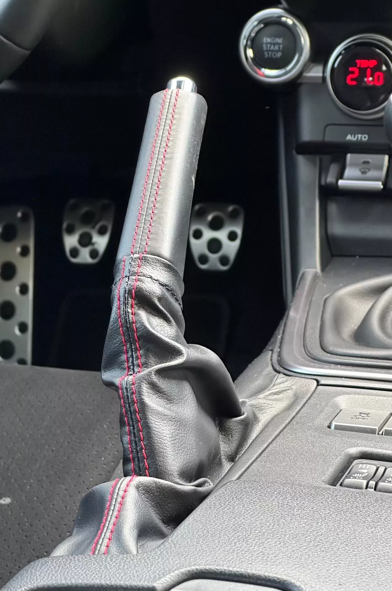 Vista detalhada do console central da Subaru BRZ 2023 destacando a alavanca de câmbio metálica com costura vermelha contrastante na bota de couro, mostrador digital exibindo '21', pedais esportivos de alumínio com furos perfurados e características tecnológicas como botões de controle de estabilidade eletrônica.