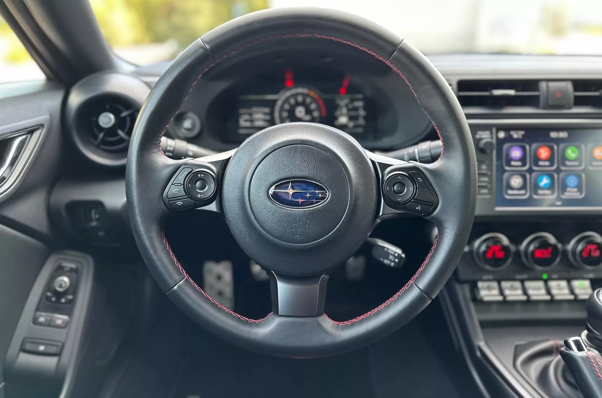 Vista interior do Subaru BRZ 2023 do assento do motorista focada no volante de três raios com costura vermelha, logo da Subaru, e painel de instrumentos com cluster de instrumentos e console central