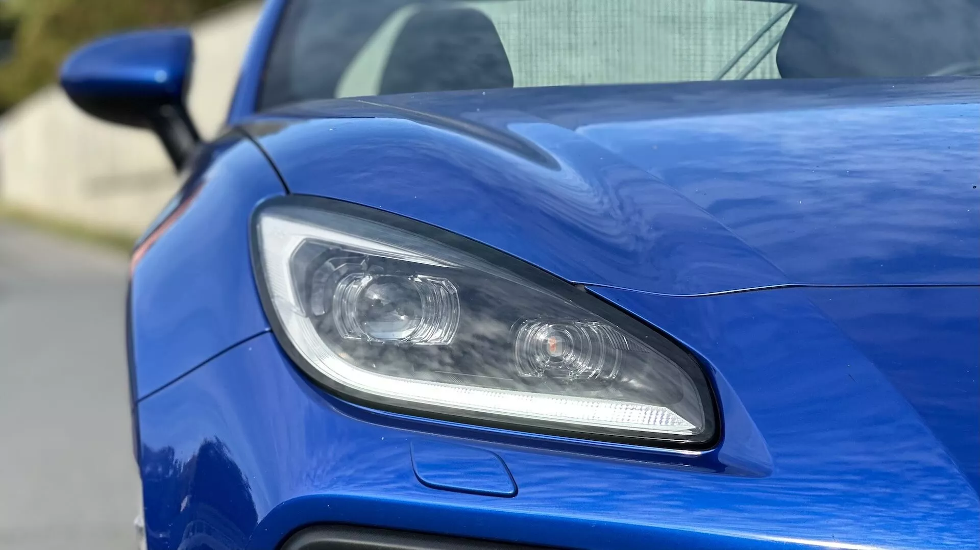 Visão detalhada do farol do lado do motorista do Subaru BRZ 2023, com iluminação LED ou HID, lentes de projetor, e entrada de ar esportiva contra uma carroceria azul metálica