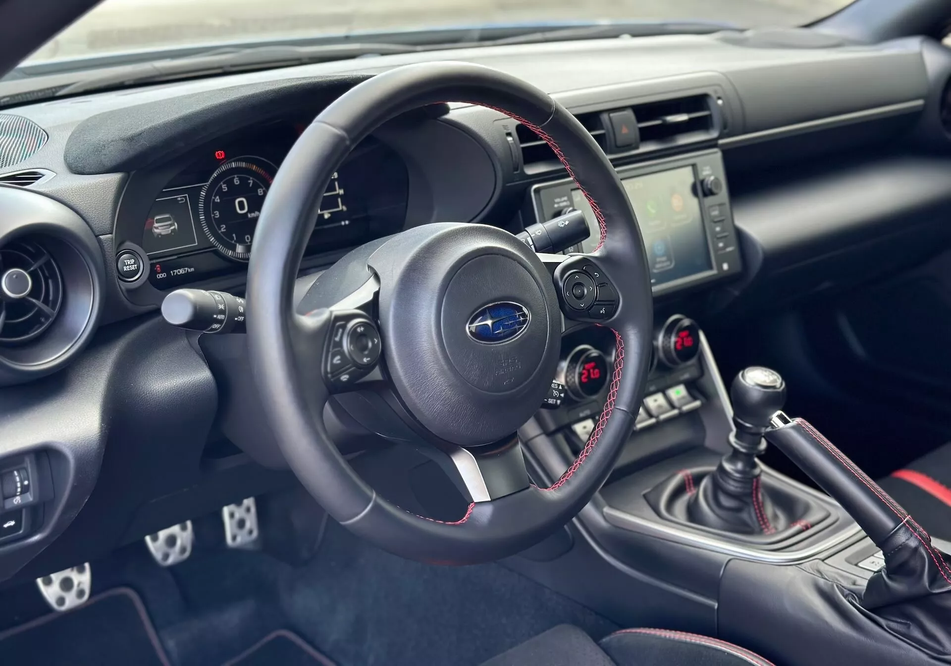 Interior do Subaru BRZ 2023 focado no lado do condutor do painel e cockpit com vista detalhada do volante, painel de instrumentos, consola central com controles de áudio e clima, e alavanca de câmbio manual com costura vermelha esportiva
