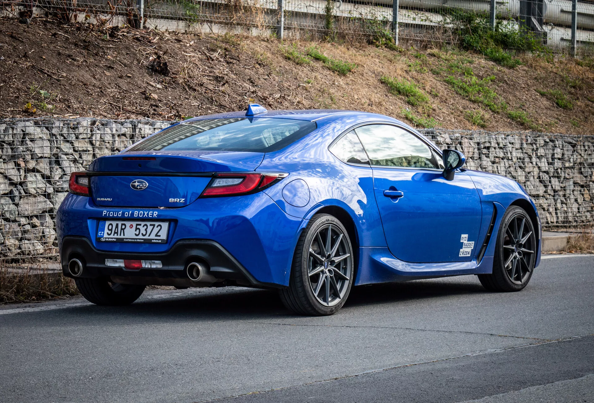 Coupé desportivo Subaru BRZ 2023 em azul metálico estacionado numa inclinação, destacando sua vista traseira três quartos que enfatiza o perfil lateral do veículo, a curvatura traseira, rodas de liga leve esportivas, pontas de escape duplas, spoiler de lábio e emblema do motor 'Proud of Boxer'.