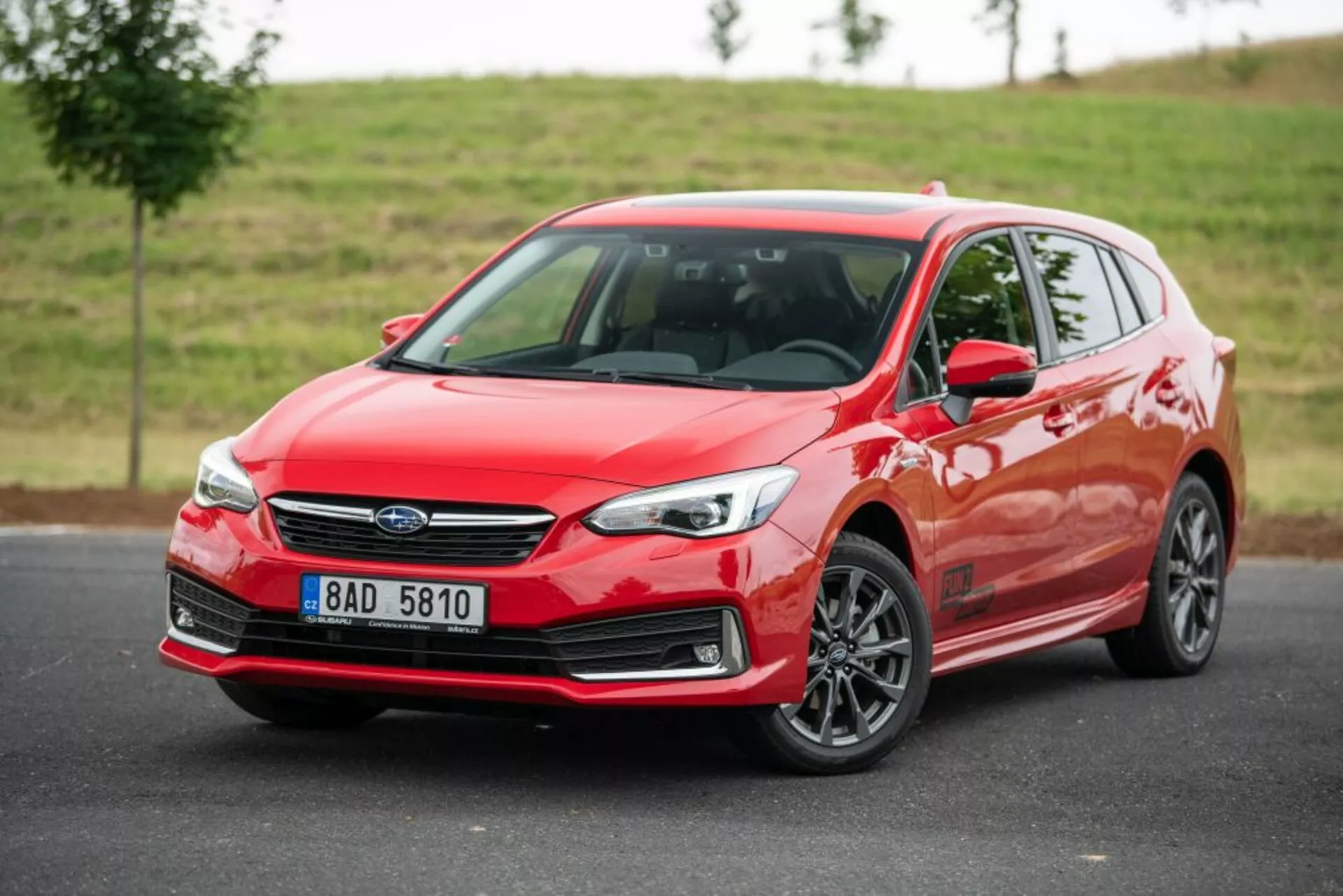 Subaru Impreza hatchback vermelho brilhante de 2018 com vista frontal de três quartos, mostrando os seus faróis LED elegantes, grade distintiva com emblema central, e rodas de liga leve multiraiadas contra um cenário natural nublado