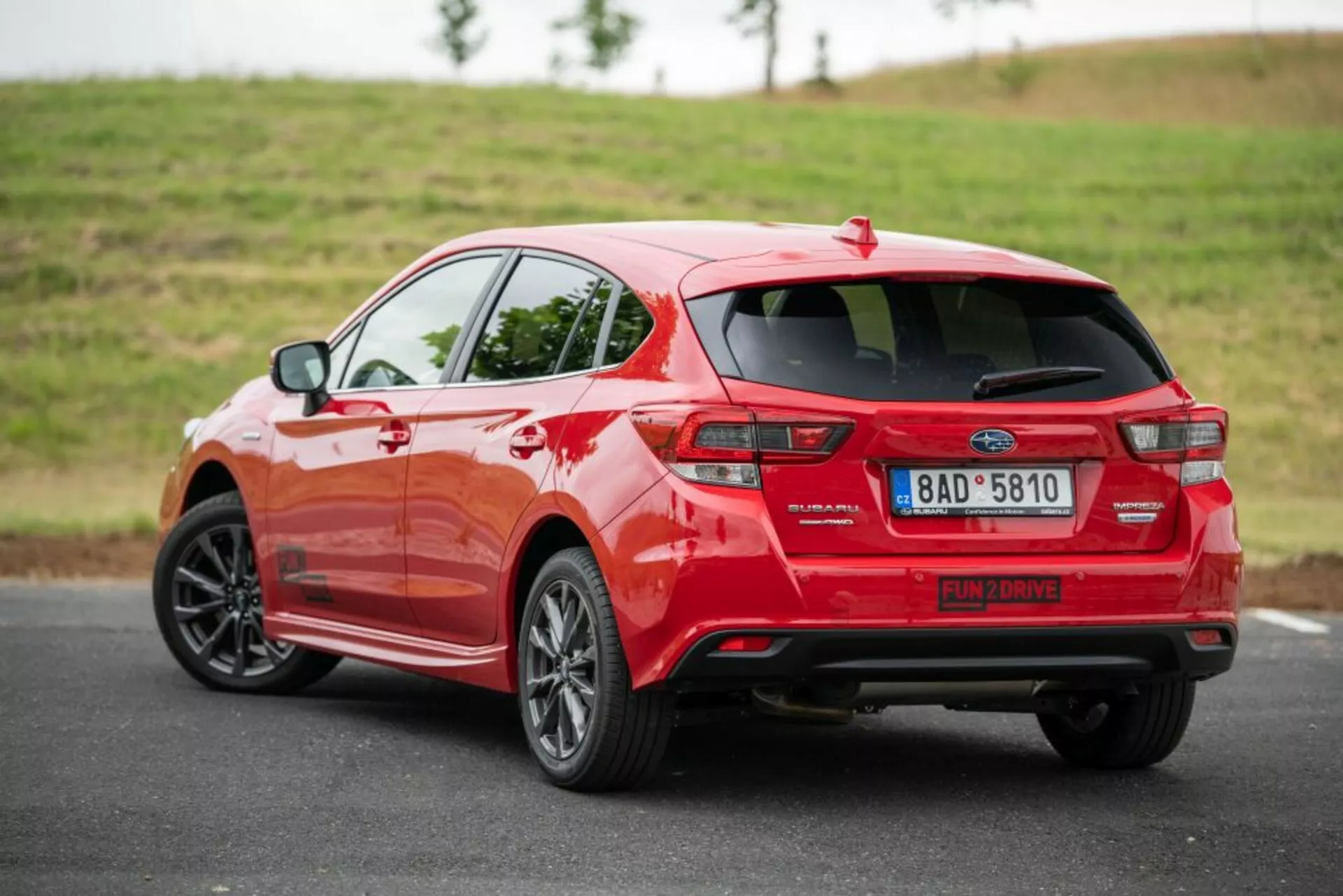 Hatchback vermelho Subaru Impreza 2018 visto de um ângulo traseiro de três quartos, destacando seu perfil lateral, design distintivo da luz traseira, sistema de escape, spoiler e rodas de liga leve em uma estrada de asfalto com um fundo gramado em iluminação suave e nublada