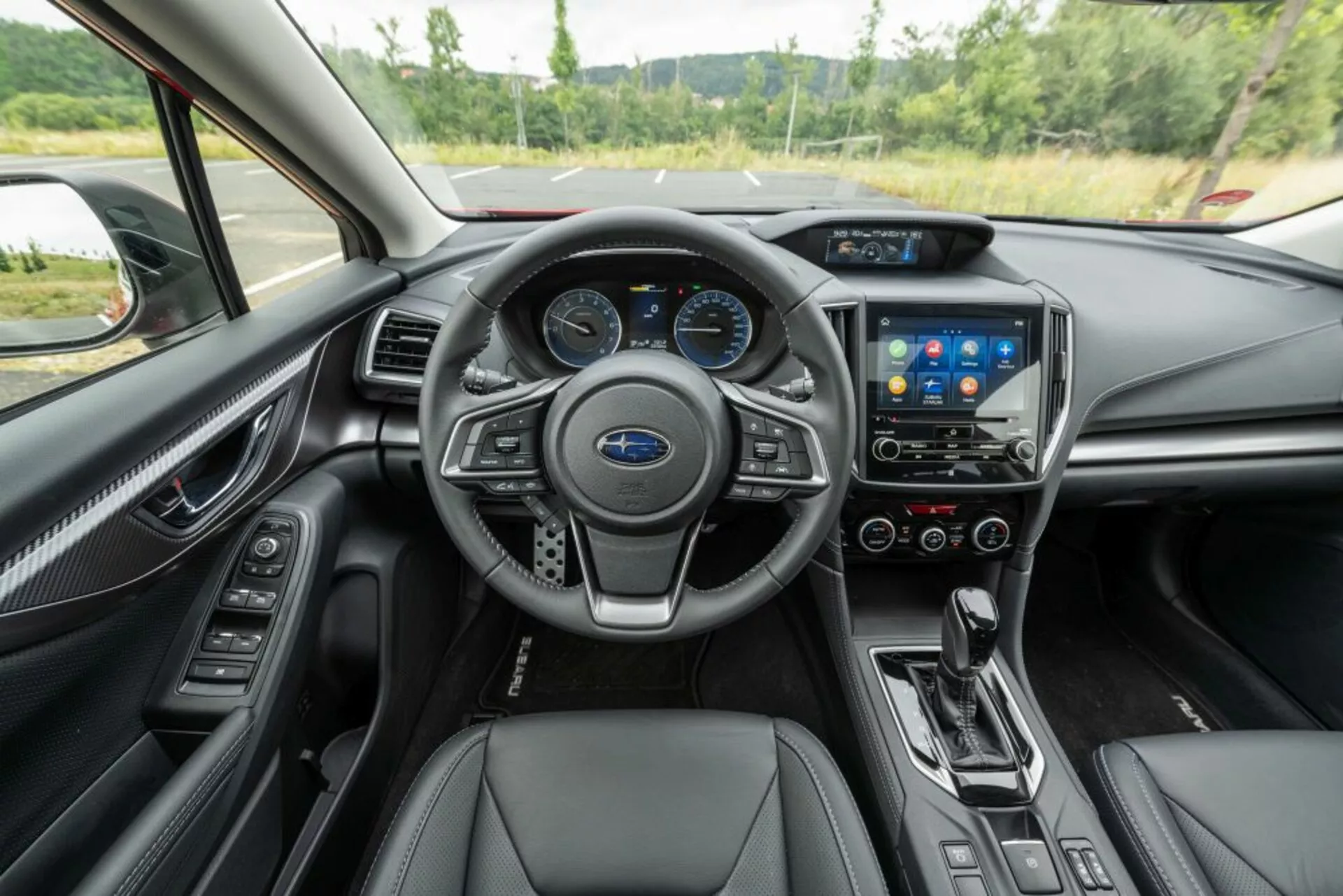 Vista interior do Subaru Impreza 2018 a partir do banco traseiro mostrando o volante de três raios revestido em couro com controles montados, emblema da Subaru visível, conjunto de instrumentos, sistema de infotainment touchscreen, configurações de controle de áudio e clima, sistema de clima de zona dupla, seletor de engrenagem de transmissão automática, assentos em couro, acabamento do painel semelhante a fibra de carbono e detalhes em metal escovado.