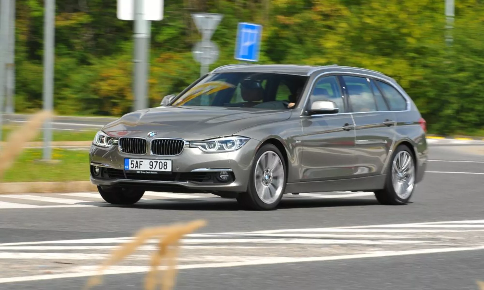 Vista dinâmica de três quartos frente de uma station wagon BMW prata em movimento com grelha e emblema distintos em forma de rim, capturada de um ângulo ligeiramente elevado indicando velocidade e movimento, foco em características de design e tecnologia como faróis angulares, rodas de liga e barras no tejadilho num ambiente externo com desfoque de movimento.