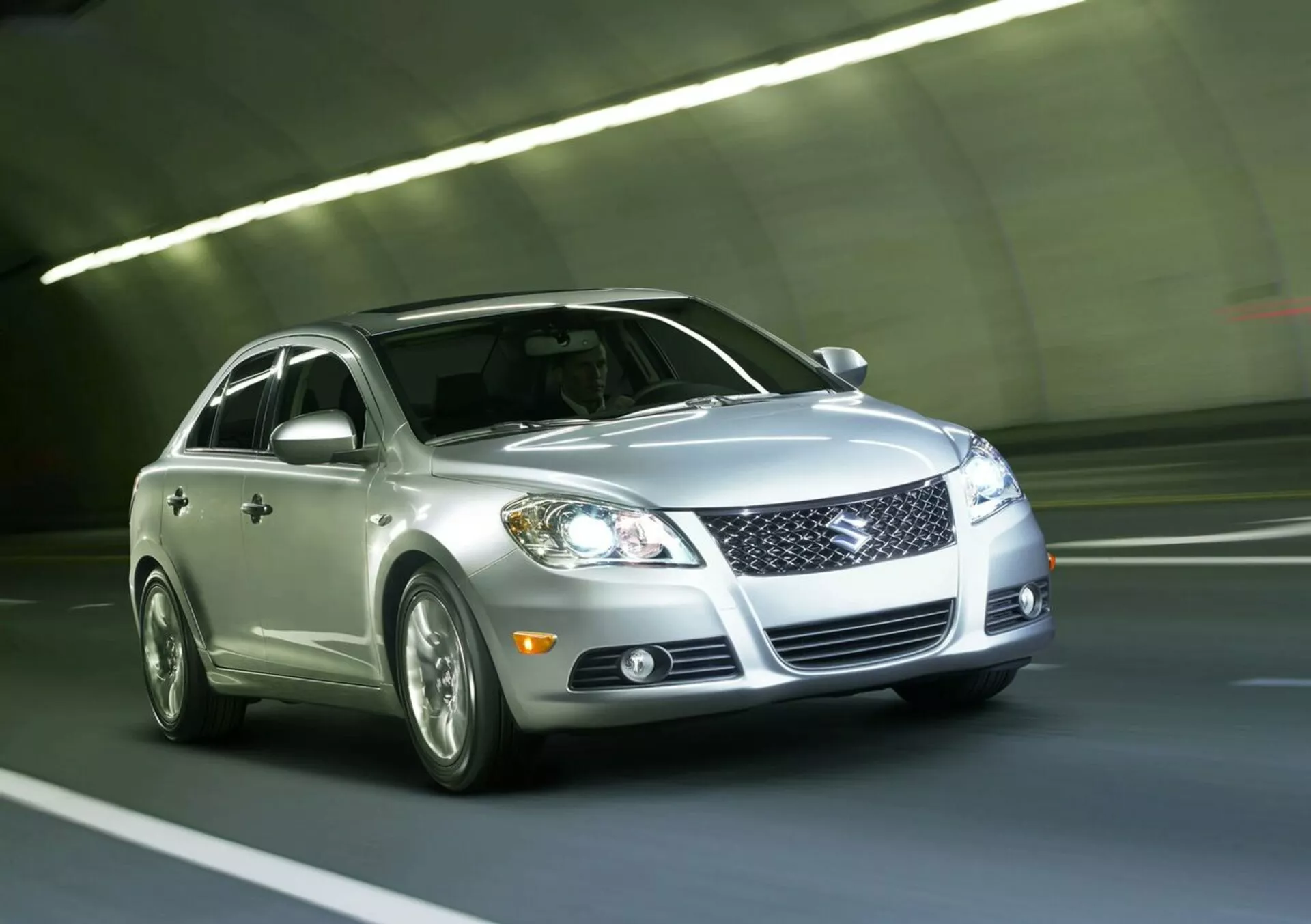 Sedan prateado Suzuki Kizashi 2010 dirigindo através de um túnel, apresentando grade frontal, faróis LED, luzes de nevoeiro, rodas de liga leve e pintura reflexiva, visto de um ângulo frontal baixo de três quartos enfatizando o design dinâmico e a tecnologia do veículo.