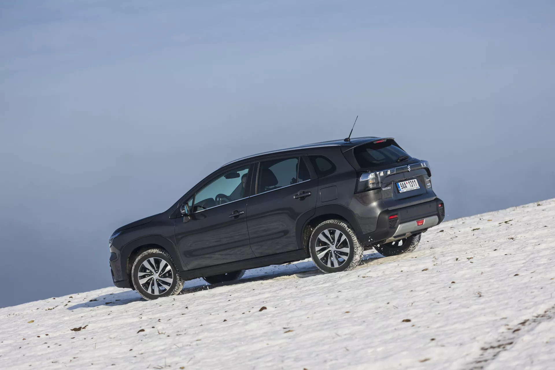 SUV Suzuki S-Cross 2022 em tonalidade escura posicionado numa encosta nevada captado de um ângulo traseiro oblíquo destacando seu perfil lateral e traseiro, grandes rodas de liga leve, janelas tintadas com desembaçador, luzes traseiras LED, aerofólio de teto, e capacidades potenciais fora de estrada sob condições de céu claro.