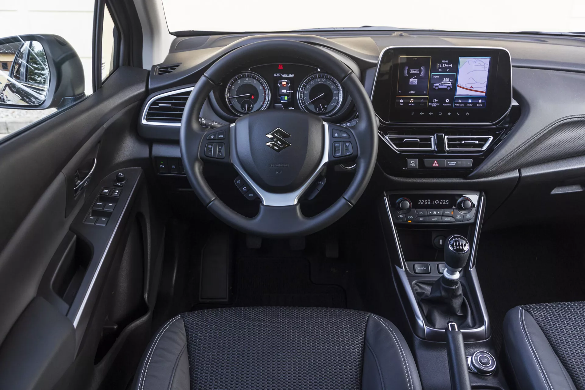 Interior da cabine frontal do Suzuki S-Cross 2022 mostrando o painel de instrumentos, volante com controles montados, cluster de instrumentos digital-analógico, sistema de infotenimento touchscreen, controles do sistema de climatização, alavanca de câmbio em couro, seletor de modo de condução, freio de estacionamento eletrônico e painel da porta com botões de controle