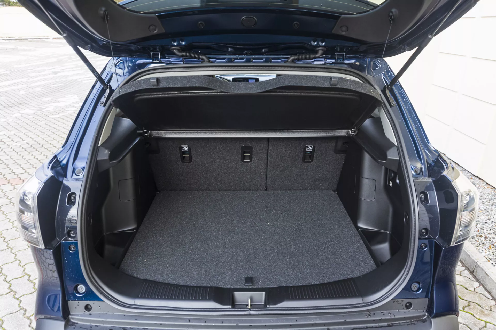Vista do espaço do porta-malas traseiro do Suzuki S-Cross 2022 com a tampa aberta mostrando uma área carpetada vazia, compartimentos laterais e bancos traseiros rebatidos
