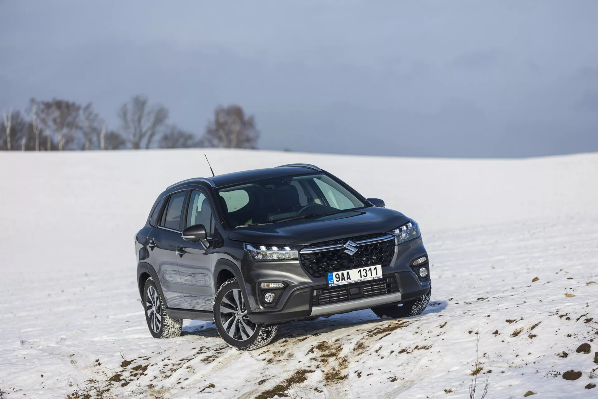 SUV compacto Suzuki S-Cross de cor escura, modelo 2022, estacionado numa paisagem nevada, apresentando uma vista frontal e lateral a um ângulo de 45 graus, exibindo uma grelha elegante com acabamento metálico, faróis de LED alongados, rodas de liga leve multirraios, e frisos das janelas cromados polidos indicando um pacote de luxo e potencial capacidade fora de estrada