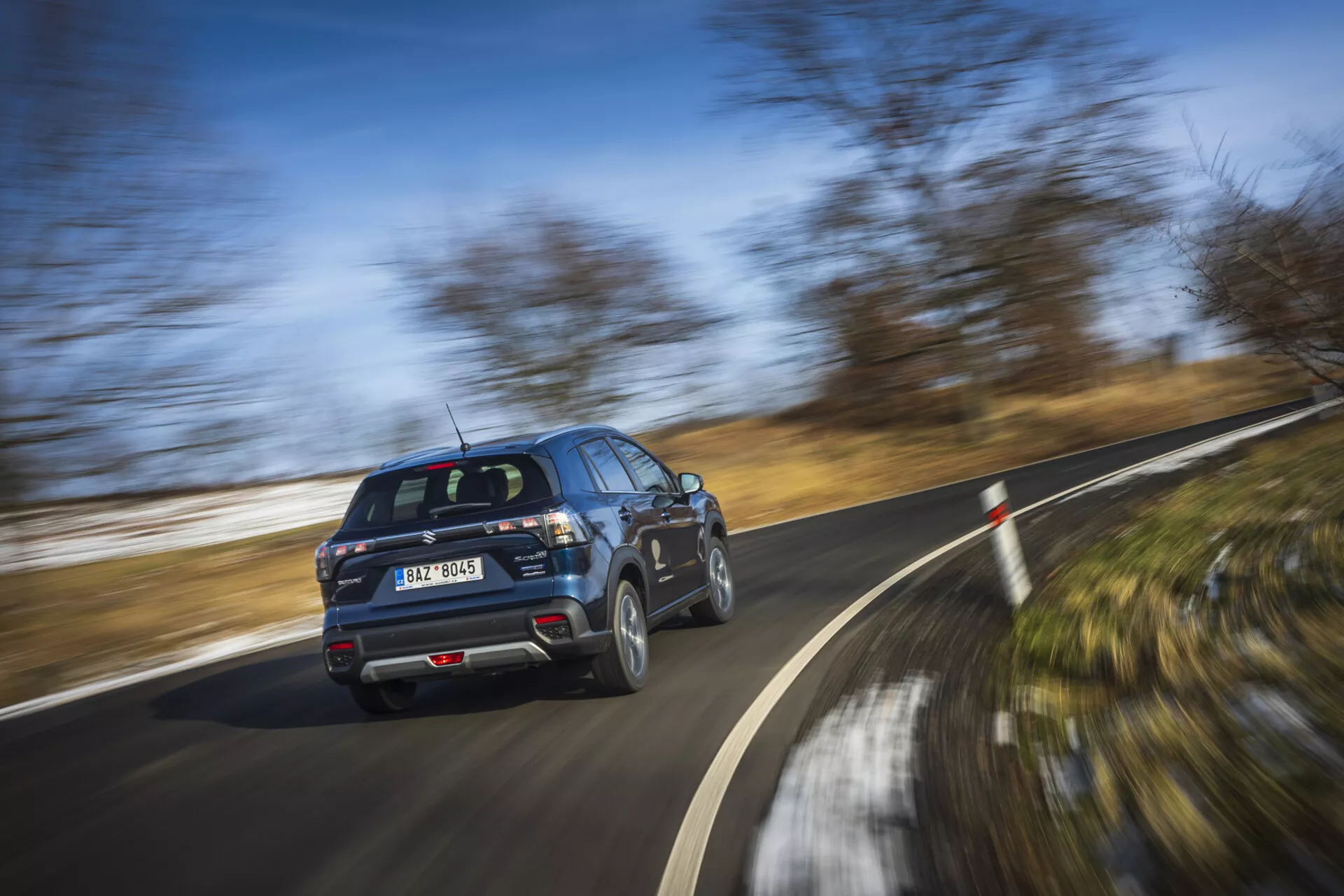 SUV moderno Suzuki S-Cross 2022 em movimento, exibindo perfil de vista traseira de três quartos com rodas girando em um dia claro