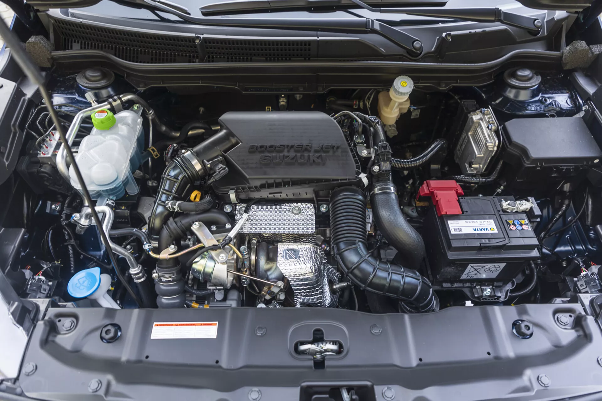 Compartimento do motor do Suzuki S-Cross 2022 mostrando uma vista de frente com componentes visíveis como o bloco do motor, a cabeça do cilindro, o coletor de admissão e características tecnológicas incluindo turboalimentador e temporização variável de válvulas, juntamente com a marca Suzuki na cobertura do motor, em condição impecável.
