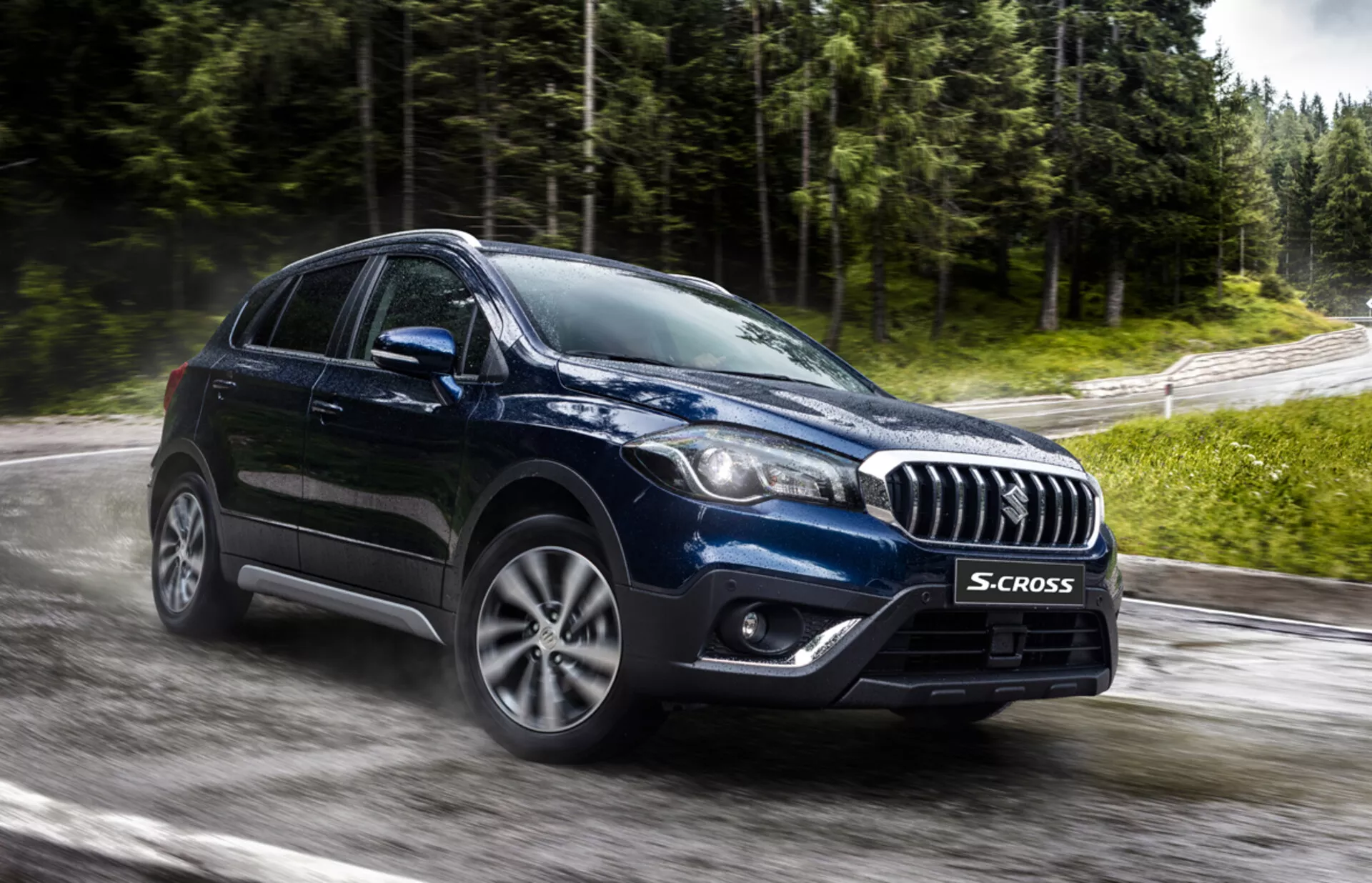 Suzuki S-Cross 2022 capturado de um ângulo frontal de três quartos destacando sua grade com detalhes em cromo, faróis modernos de LED ou HID, rodas de liga leve multiraiadas e forma robusta de crossover em uma estrada molhada e cercada de verde, sugerindo tecnologia avançada e capacidade para todo tipo de clima.