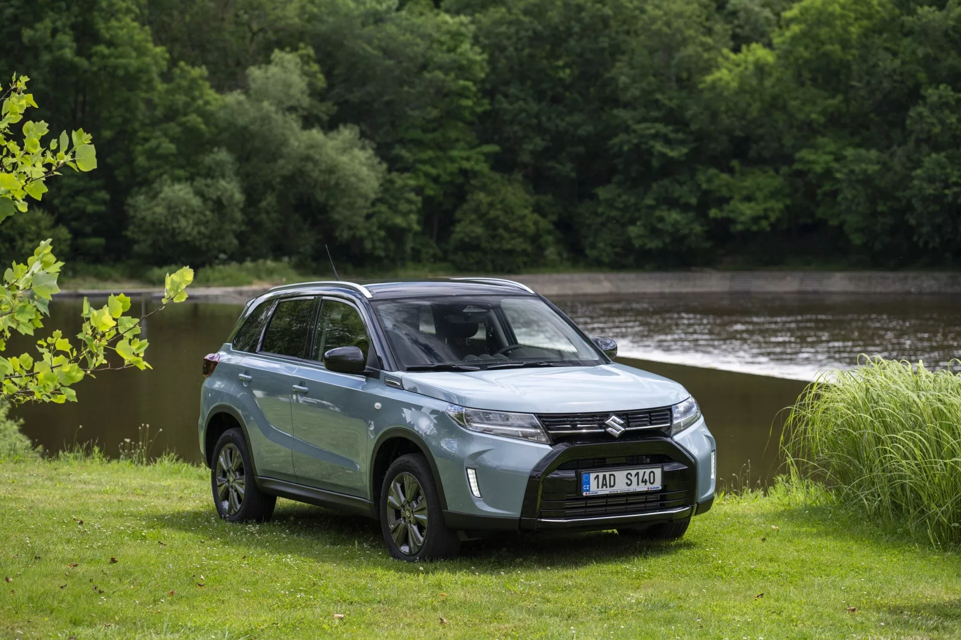 Fotografia de um SUV moderno Suzuki Vitara 2024 (2ª geração, facelift 2) com carroceria em dois tons azul-claro com teto preto, estacionado em um campo de grama com água e árvores ao fundo. O SUV, visto de um ângulo frontal-quart mostrando o lado do motorista, apresenta uma grade frontal proeminente com um emblema central, faróis em LED angulares elegantes, capô liso, rodas de liga leve multirraiadas e distância ao solo notável. As janelas escurecidas, os espelhos laterais e as maçanetas das portas da cor da carroceria destacam seu design contemporâneo, enquanto os espelhos laterais integram luzes indicadoras LED.