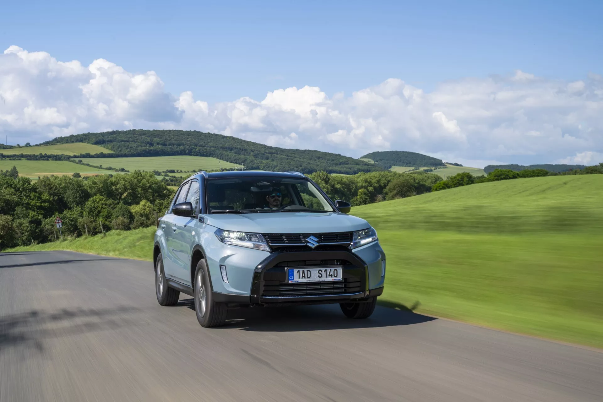 Foto de ângulo baixo do facelift da segunda geração do Suzuki Vitara 2024 conduzindo em uma estrada, vista de três quartos frontal mostrando a grade, os faróis e o desfoque de movimento em um cenário verde e montanhoso.