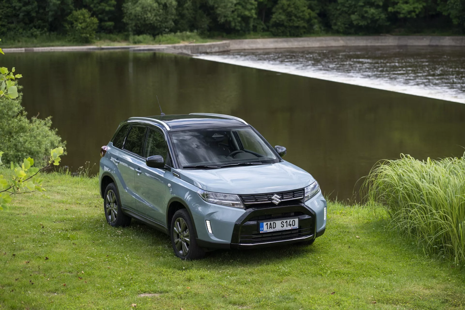 A imagem mostra o SUV Suzuki Vitara 2024 (segunda geração, segundo facelift) em um ambiente externo sereno perto de um corpo d'água. Capturada de um ângulo frontal de três quartos, os faróis LED elegantes, a grade e o para-choque dianteiro do SUV são claramente visíveis. O capô esculpido e o para-brisa inclinado contribuem para o perfil aerodinâmico. Um lado do SUV revela seu design de quatro portas, área de carga traseira fechada, rodas de liga leve e pneus robustos para todos os tipos de terreno. A cor prata ou cinza suave acrescenta um toque refinado. O terreno natural e não pavimentado ao redor do SUV sublinha suas capacidades off-road e seu apelo aventureiro.
