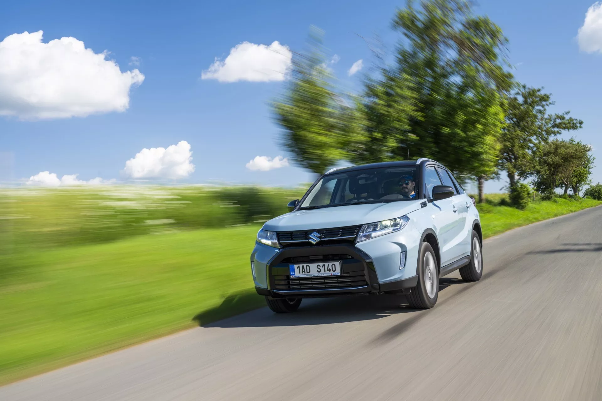 Imagem dinâmica do Suzuki Vitara 2024 (2ª geração, facelift) capturada de uma visão frontal de três quartos. O carro está circulando em uma estrada com uma paisagem verde desfocada ao fundo, sugerindo movimento. Características visíveis incluem o capô, a grade, os faróis, o para-choque dianteiro e uma roda dianteira ligeiramente girada. O lado do motorista, incluindo a porta e o retrovisor, é claramente visível. O desfoque de movimento e as listras ao longo do ambiente aumentam a sensação de velocidade enquanto o carro permanece em foco. Linhas de contorno, cortes laterais e o design da roda são evidentes. Condições de luz diurna brilhante com céu claro e sombras sugerem luz solar vindo do canto superior esquerdo. Pode-se ver uma pessoa através do para-brisa, provavelmente o motorista.