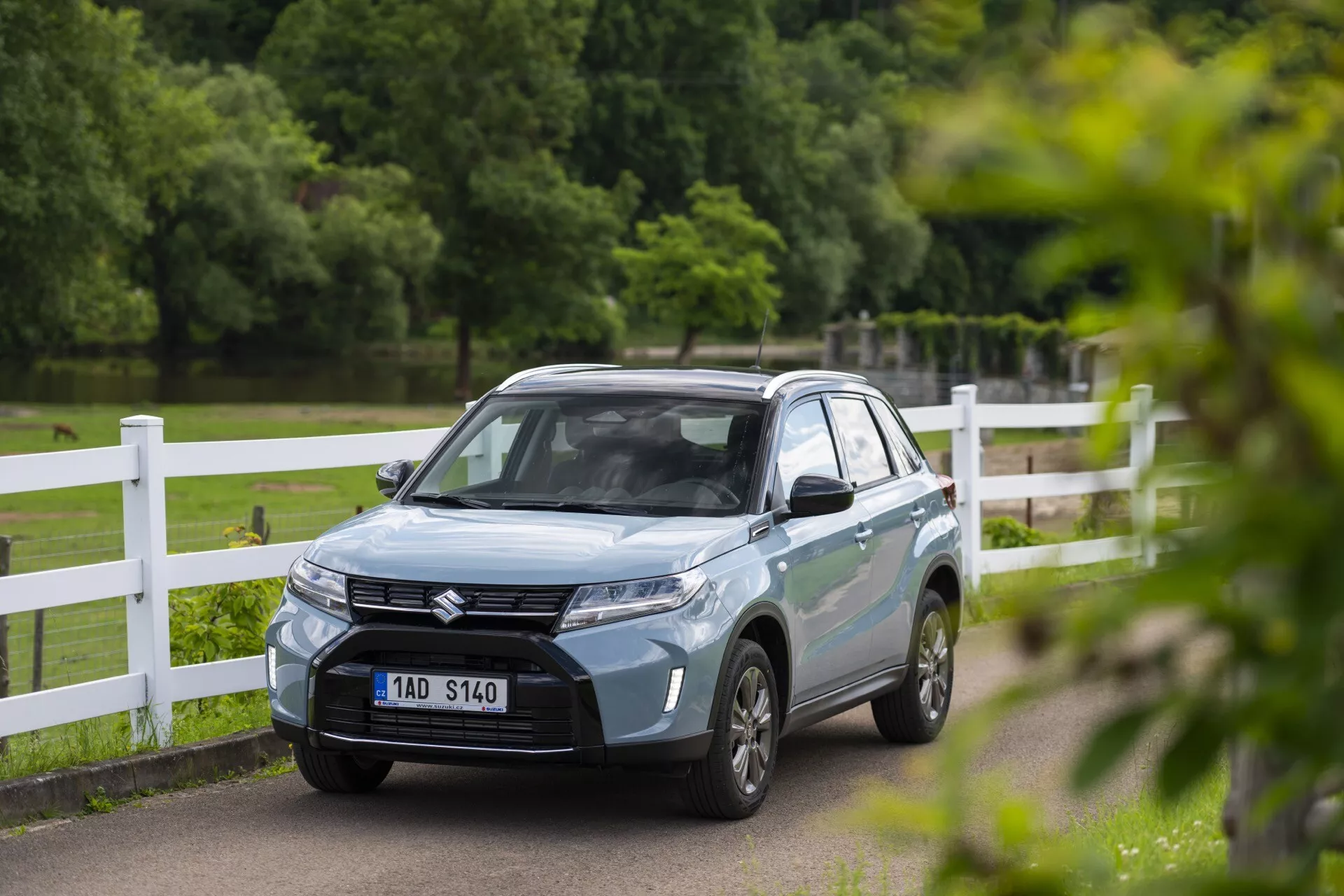 SUV Suzuki Vitara 2024 azul claro em um cenário de parque ou campo, visto de um ângulo de três quartos frontal que mostra sua grade sofisticada com emblema, faróis LED afiadas, linhas aerodinâmicas do capô e rodas de liga leve multi-raios. A placa frontal do veículo está desfocada e os espelhos retrovisores podem ter indicadores de direção integrados. A foto captura o carro com pouca profundidade de campo, enfatizando seu design moderno enquanto o fundo permanece ligeiramente desfocado.