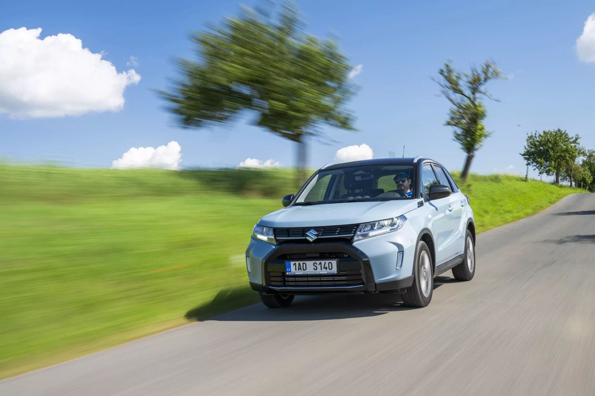 Fotografia dinâmica do Suzuki Vitara 2024 (segunda geração facelift), exibindo seu design moderno e velocidade. Capturado de uma perspectiva de quarto dianteiro esquerdo, a imagem destaca os faróis nítidos, a grade frontal proeminente e as rodas de liga padrão. O efeito de desfoque de movimento no fundo e nas rodas acentua o movimento rápido do carro, visto em uma estrada borrada com árvores e grama.