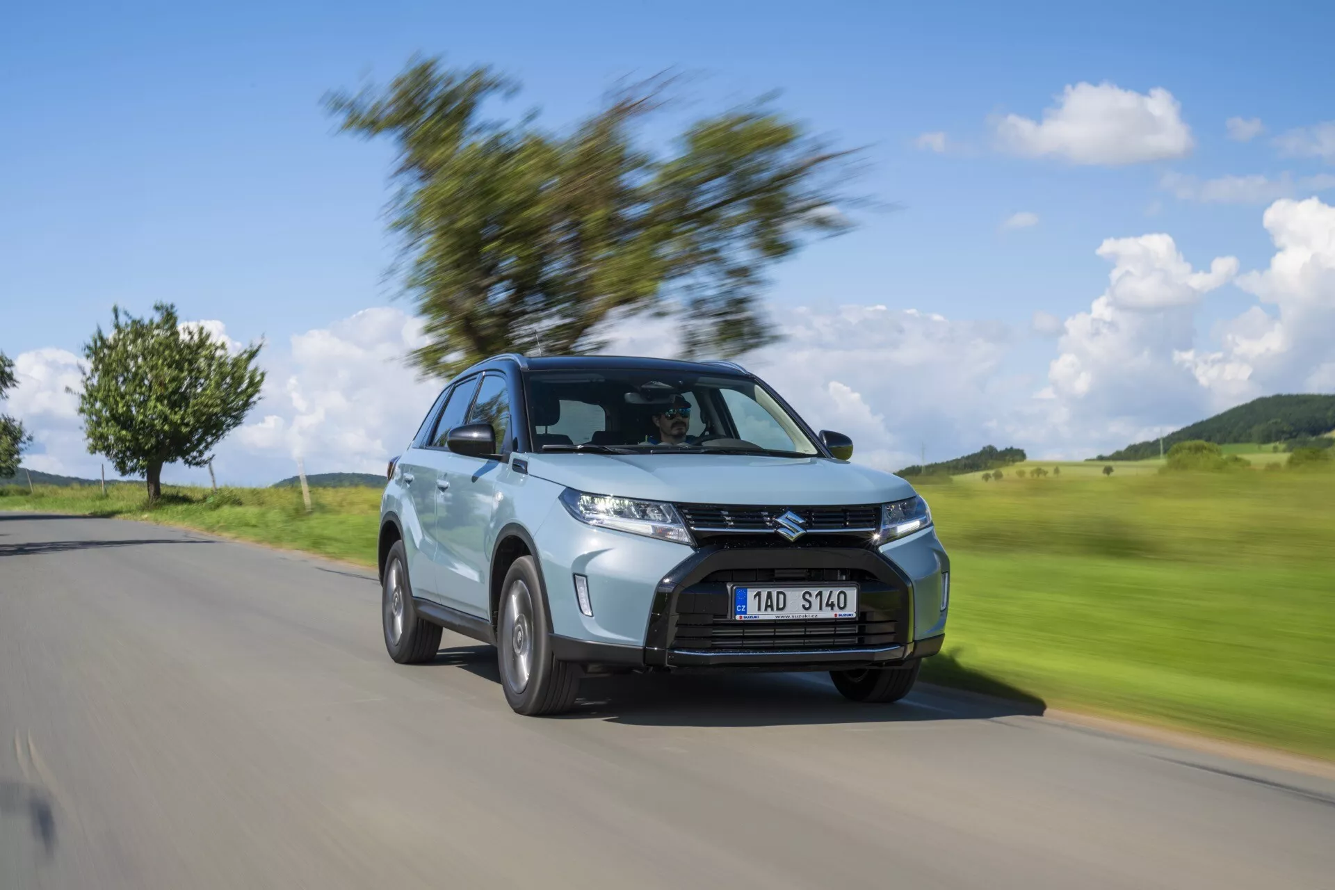 Imagem dinâmica do Suzuki Vitara 2024 (2ª geração, facelift 2) em movimento, mostrando o ângulo de três quartos frontais. O carro está em foco nítido, enquanto o fundo está desfocado pelo movimento, destacando seus faróis, grade proeminente, rodas de liga leve, carroceria SUV compacta, pintura metálica e design aerodinâmico. Ideal para mostras automotivas e uso promocional.