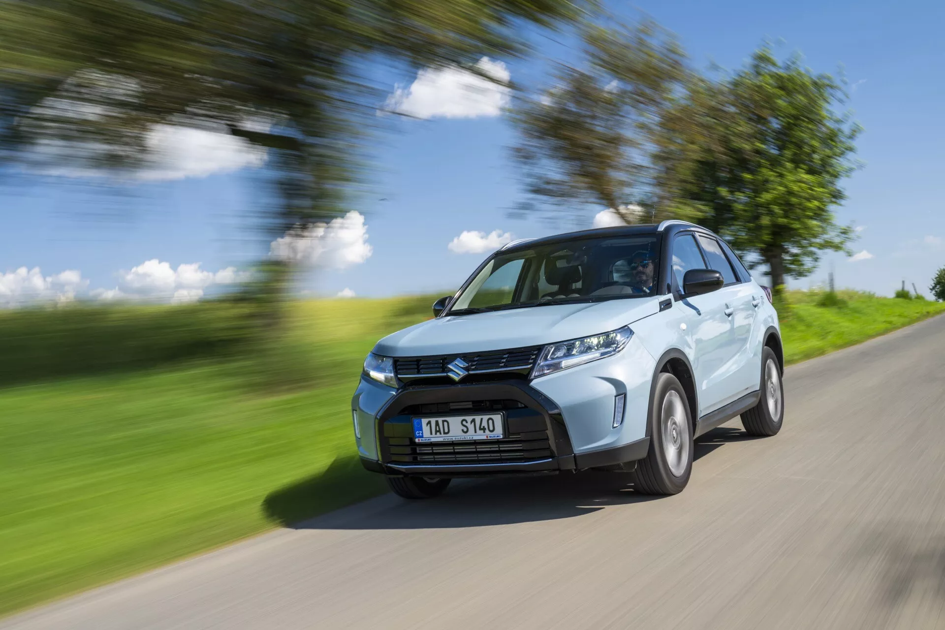 Um SUV compacto Suzuki Vitara 2024 (2ª geração facelift) azul claro em movimento numa estrada rural. A imagem captura o quarto dianteiro do carro, mostrando a grelha frontal, faróis, parte do capô, para-brisas e lado do motorista, incluindo o arco da roda dianteira, parte da porta traseira e uma porção do arco da roda traseira. O carro está nítido contra um fundo desfocado e a estrada, indicando velocidade. As rodas têm um ligeiro desfoque sugerindo rotação. Iluminado por luz natural brilhante num dia ensolarado, lançando sombras na parte inferior do veículo, a imagem destaca elementos de design como os vincos no capô, o design do para-choques dianteiro e o padrão da grelha. Os faróis LED são visíveis, potencialmente indicando características modernas como sensores de estacionamento. A placa de matrícula é visível sem informações sensíveis.