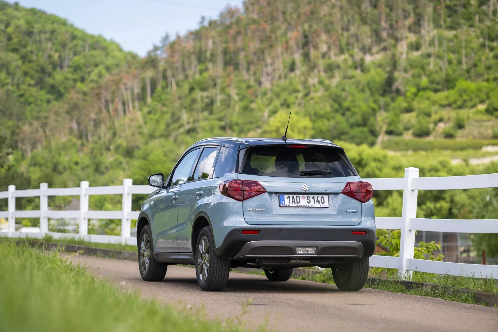 A imagem mostra o Suzuki Vitara 2024 (2ª geração, facelift) de um ângulo traseiro de três quartos, destacando seu perfil lateral direito traseiro enquanto está posicionado em uma estrada e ligeiramente inclinado para a esquerda. Características visíveis incluem a porta traseira, o para-brisa traseiro, portas e janelas parciais do lado direito do passageiro, luzes traseiras de LED, sensores de estacionamento no para-choque traseiro, spoiler de teto e uma antena curta no teto. O carro tem rodas de liga e uma placa de registro visível. Está situado contra um pano de fundo rural com uma cerca branca e vegetação exuberante.