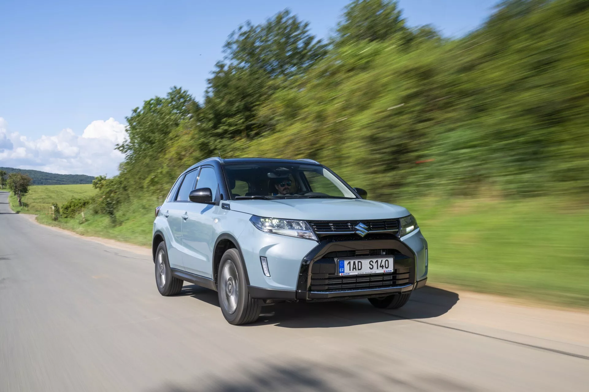 Um SUV moderno, o Suzuki Vitara 2024 (segunda geração, facelift 2) mostrado em movimento de um ângulo baixo e três quartos da frente. A imagem destaca a distinta dianteira do veículo, incluindo uma grade proeminente com ripas horizontais, faróis LED afiados e angulares, e uma roda de liga leve multi-raios. O SUV é capturado dirigindo em uma estrada de asfalto com uma paisagem verde natural ao fundo, com um desfoque de movimento que transmite velocidade e dinamismo. Características adicionais incluem um capô inclinado, espelhos aerodinâmicos para melhorar a aerodinâmica, altura elevada do solo e um interior espaçoso, tudo acentuado por um acabamento de pintura metálica.