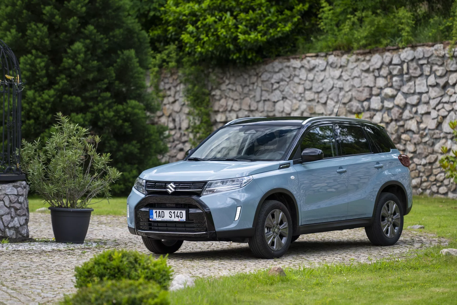 Vista frontal lateral em ângulo de um moderno SUV Suzuki Vitara 2024 estacionado ao ar livre em uma área residencial ou similar a um parque com um gramado bem cuidado e um muro de pedra ao fundo. A imagem, tirada de uma distância média, mostra a grade frontal com barras horizontais com o logotipo da empresa, faróis LED elegantes com luzes diurnas, para-choque esculpido com luzes de neblina integradas, rodas de liga, retrovisores laterais com sinais integrados, acabamento de pintura metálica, linhas dinâmicas de carroceria, vidros escurecidos, barras no teto e antena no teto, destacando seu design e características modernas.