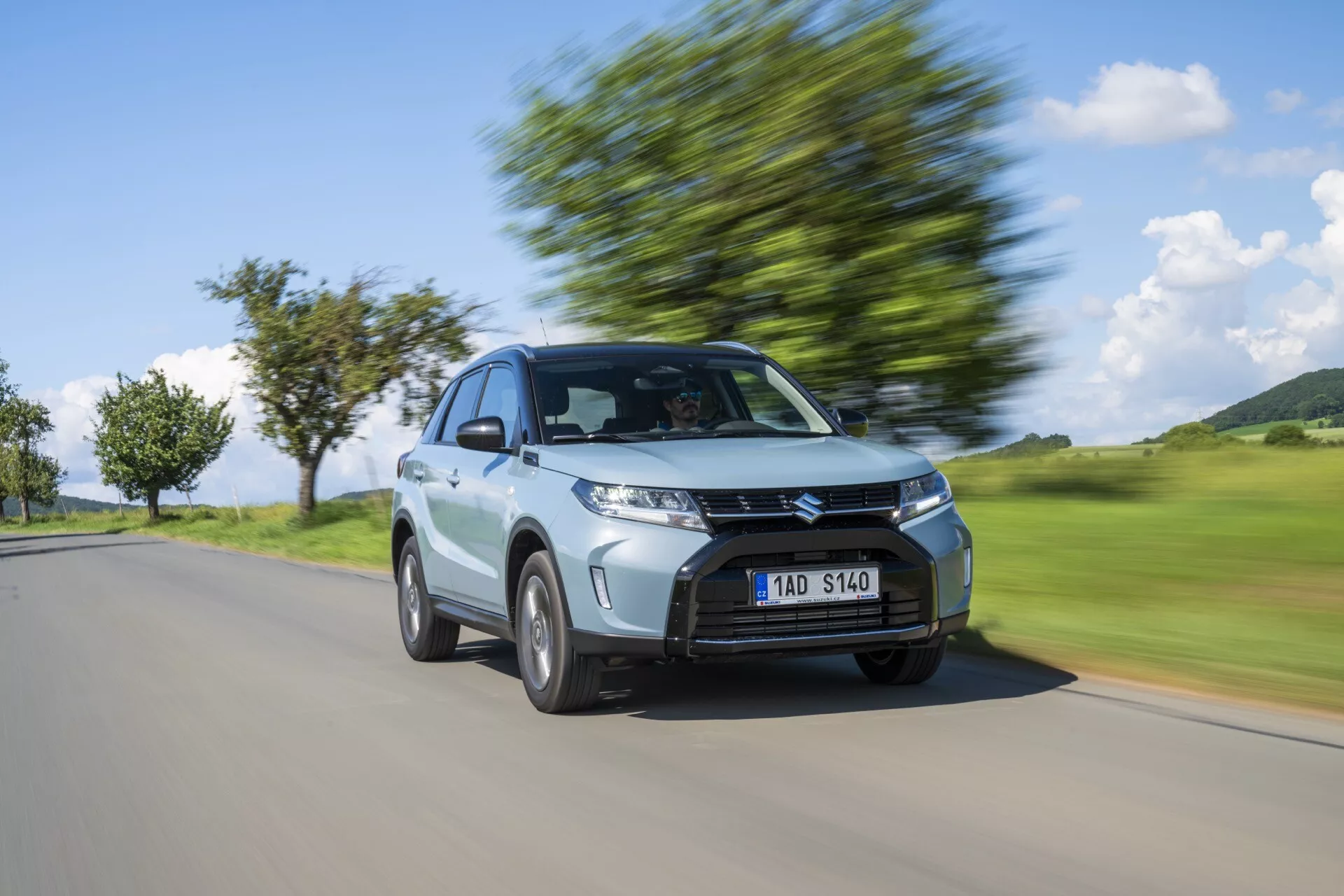 Imagem dinâmica de um carro hatchback prateado Suzuki Vitara 2024 em movimento capturada de um ângulo frontal de três quartos, destacando seu design moderno com uma grade proeminente, faróis angulares, teto inclinado e rodas de liga em movimento com fundo desfocado indicando velocidade.