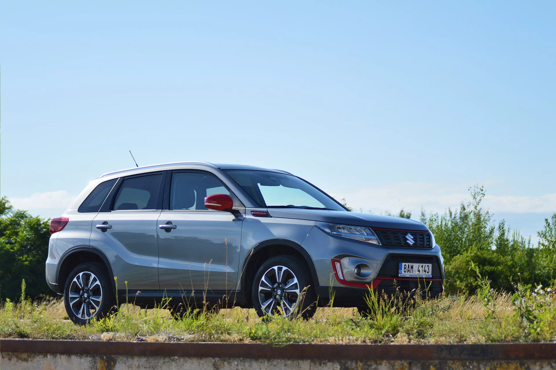 Vista de perfil lateral de um SUV compacto Suzuki Vitara 2018 de cor prateada, estacionado paralelamente a uma linha de trem num dia ensolarado. As linhas de design, as rodas de liga leve com design multirraios, o acabamento de pintura metálica, as portas e janelas fechadas, o formato dos faróis, o espelho lateral com potenciais indicadores de direção integrados e o emblema do nível de acabamento são claramente visíveis, destacando o design e as características tecnológicas do veículo.