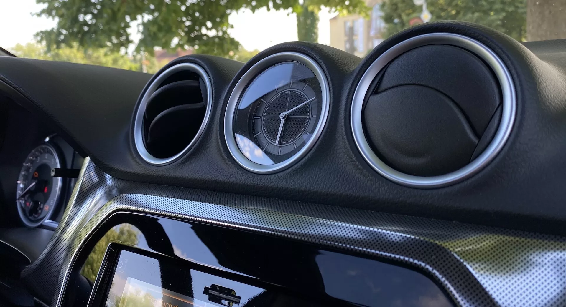 Visão interna de perto do Suzuki Vitara 2018 centrada no painel central. O painel possui três saídas de ar circulares com aparência metálica e emblema do fabricante, sistema de infoentretenimento sensível ao toque com moldura preta brilhante, material com aparência de fibra de carbono e iluminação ambiente para melhorar a estética.