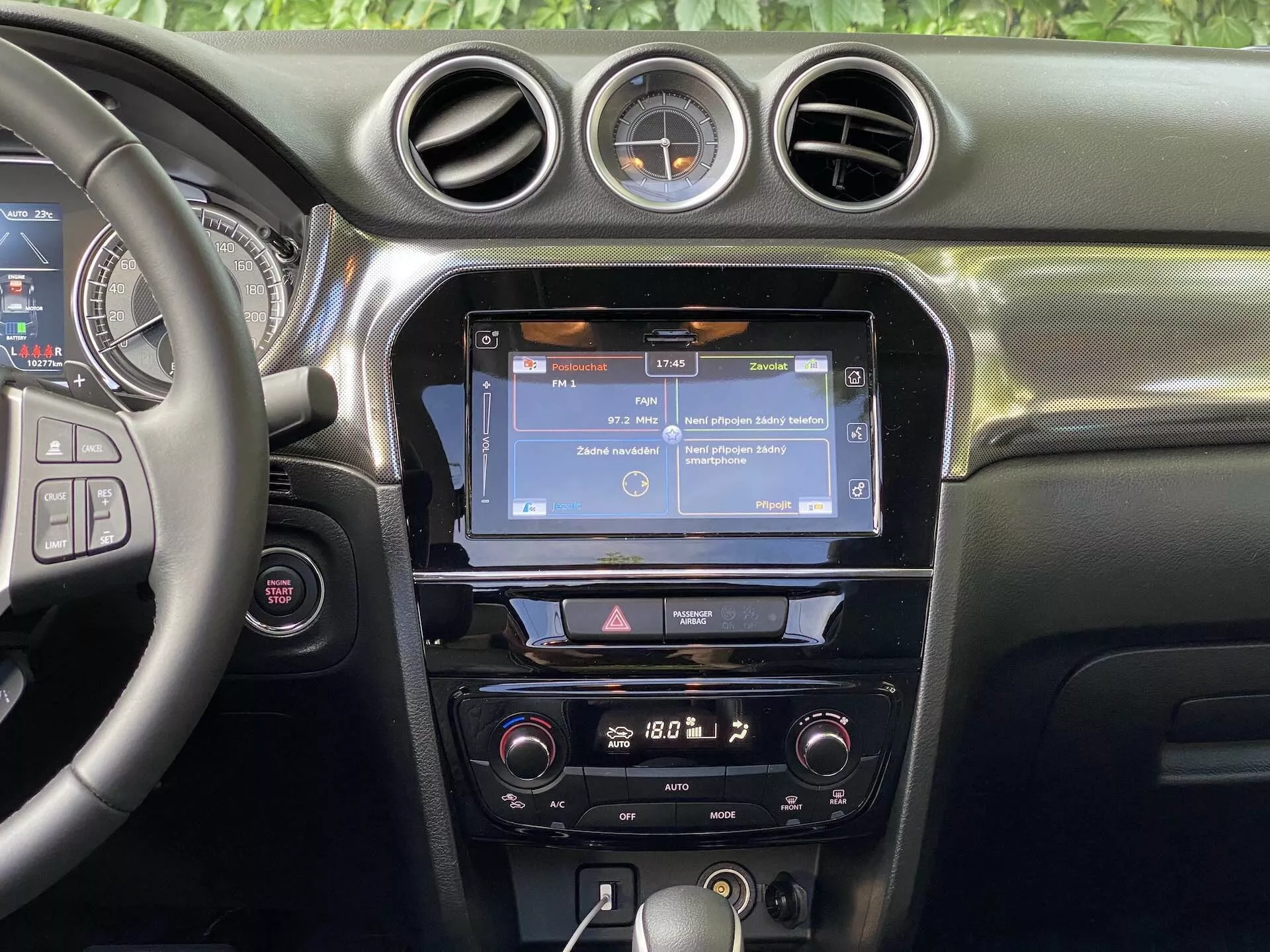 Vista interior do Suzuki Vitara 2018 focando no painel central. A seção apresenta um volante parcialmente visível à esquerda com vários botões de controle, provavelmente para áudio, chamadas viva-voz e controle de cruzeiro. A consola central exibe uma tela sensível ao toque de infoentretenimento, possivelmente mostrando navegação ou um menu, ladeada por ventiladores verticais e coroada por três ventiladores circulares. Abaixo da tela sensível ao toque, há uma fileira de botões e discadores que proporcionam acesso rápido às configurações do infoentretenimento e do veículo. Os controles de climatização estão posicionados mais abaixo, sugerindo um sistema de controle de clima de duas zonas. O painel possui um material macio ao toque com costuras visíveis, e um relógio analógico redondo está montado entre os ventiladores centrais circulares. A visibilidade está focada nas características do painel, sem janelas ou para-brisas visíveis.