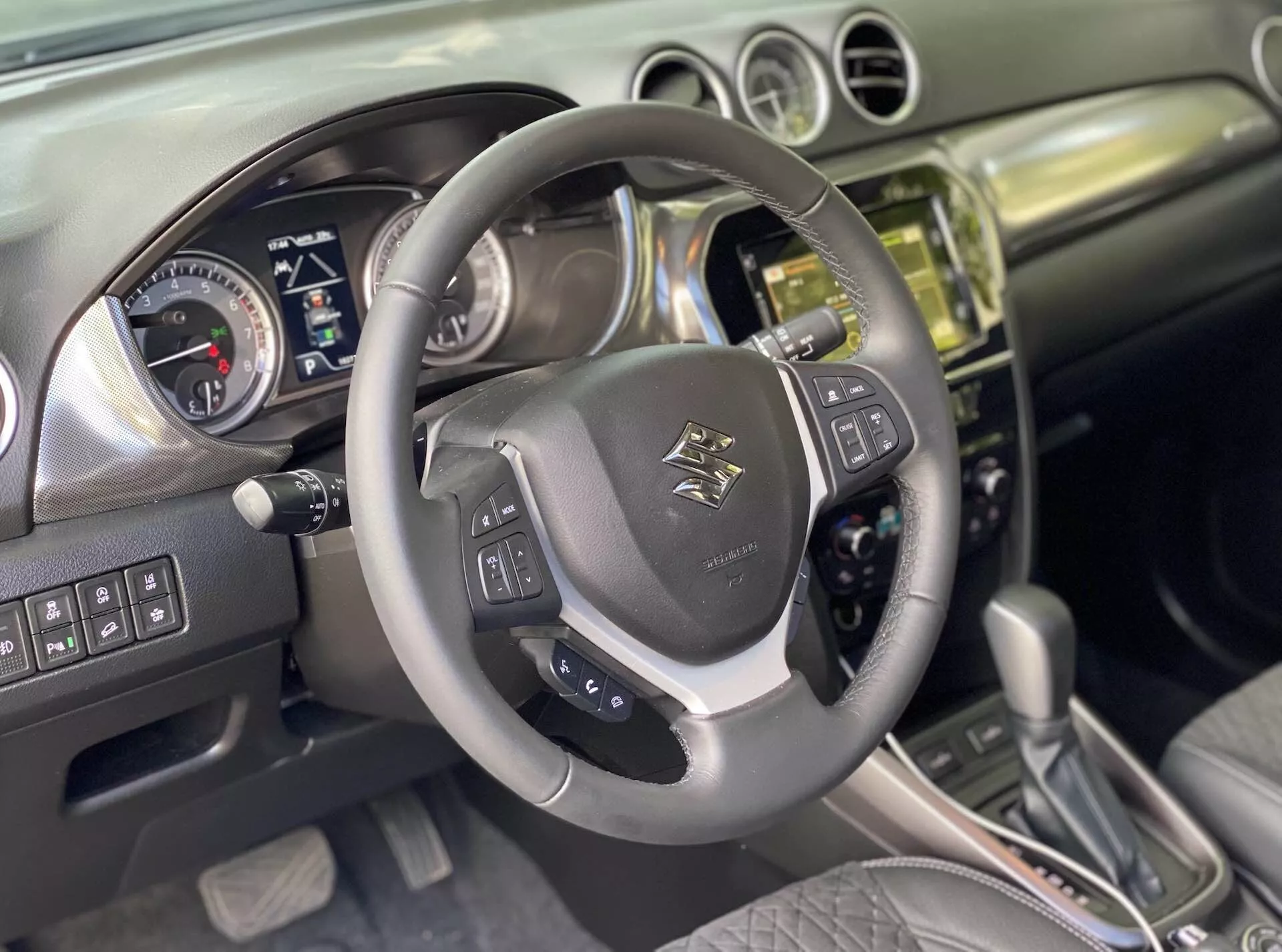 Interior do Suzuki Vitara 2018, capturando a perspectiva do motorista. Close-up de um volante multifuncional revestido em couro com emblema no centro, controles montados para áudio, controle de voz e controle de cruzeiro com detalhes metálicos. Atrás, um painel de instrumentos com mostradores analógicos para velocímetro e tacômetro, e uma tela digital para informações do veículo, sugere que o carro está desligado ou no modo acessório. A consola central exibe uma pilha com acabamento em metal ou cromado com um sistema de infotainment com tela sensível ao toque, botões e controles para o clima e o veículo. A alavanca de câmbio automática com detalhes em couro sugere vários modos de condução. O interior combina couro e metal escovado, refletindo um design moderno. Recursos avançados inferidos incluem Bluetooth, navegação, assistentes de estacionamento e sistemas de assistência ao motorista.