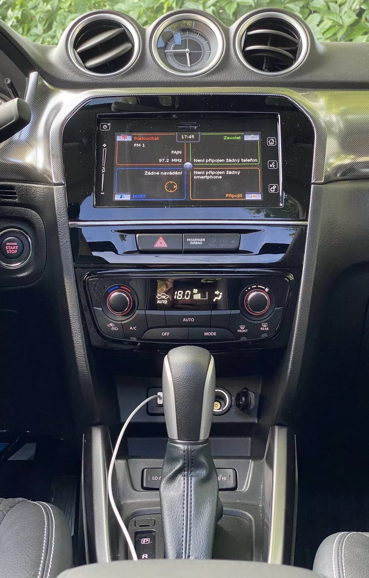 Vista detalhada do interior do Suzuki Vitara 2018, destacando o console central e o painel de instrumentos da perspectiva dos bancos dianteiros. A imagem destaca a tela sensível ao toque do sistema de infoentretenimento mostrando um menu, o sistema de controle de clima de duas zonas com leituras digitais e a alavanca de câmbio automático. O interior apresenta acabamentos em cores escuras com uma mistura de texturas foscas e brilhantes, ilustrando a qualidade do material e a tecnologia automotiva moderna.