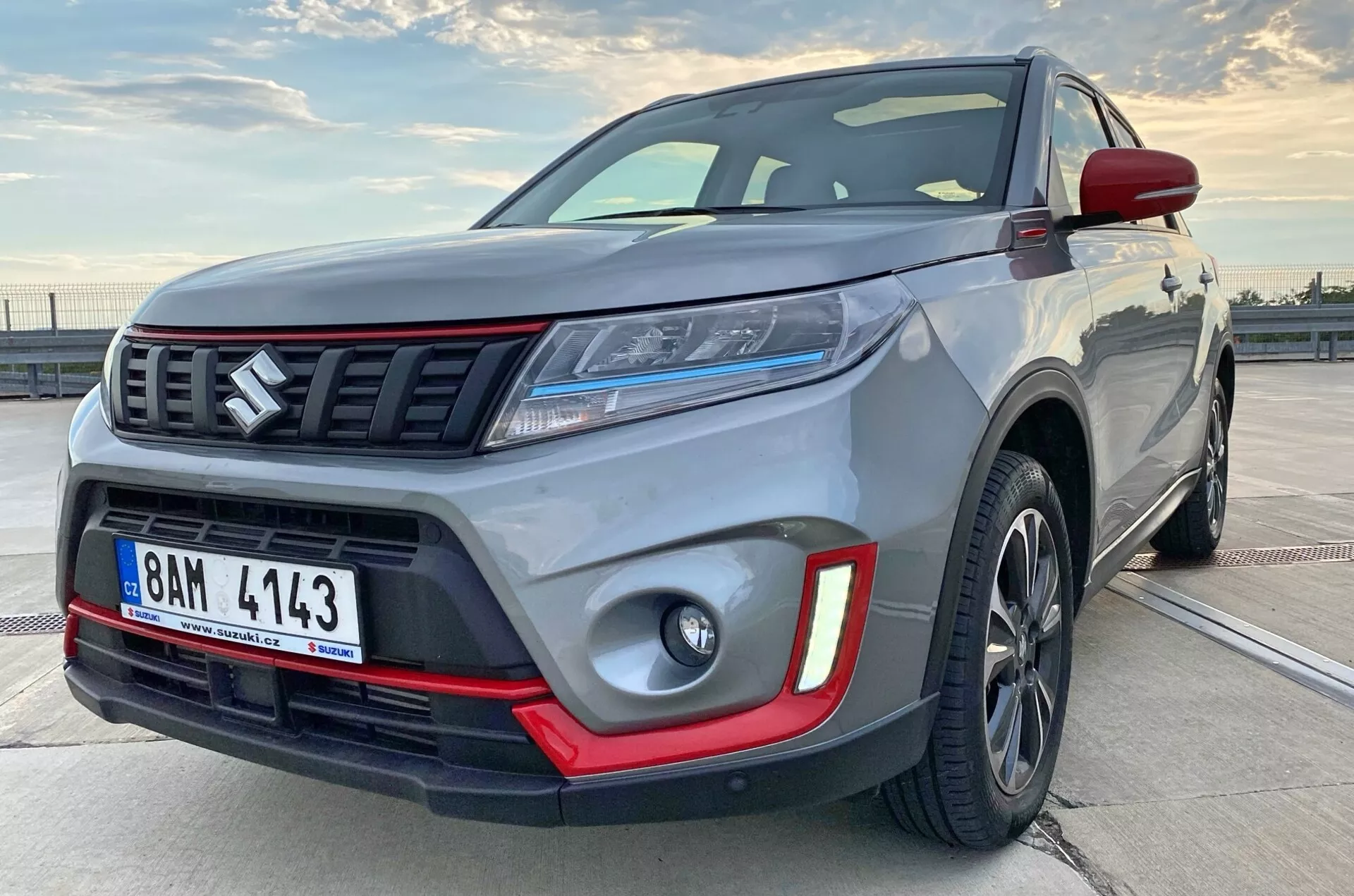 Vista lateral frontal próxima de um SUV compacto moderno, angulado para mostrar parte do perfil lateral. A imagem destaca a robustez do veículo com detalhes visíveis como o para-choque dianteiro, a grade com barras horizontais e emblema, faróis elegantes de LED, luzes de neblina com inserções vermelhas, rodas de liga e para-lamas musculosos. As rodas de liga escuras e a peça de adorno de cor contrastante sugerem uma versão premium ou esportiva.