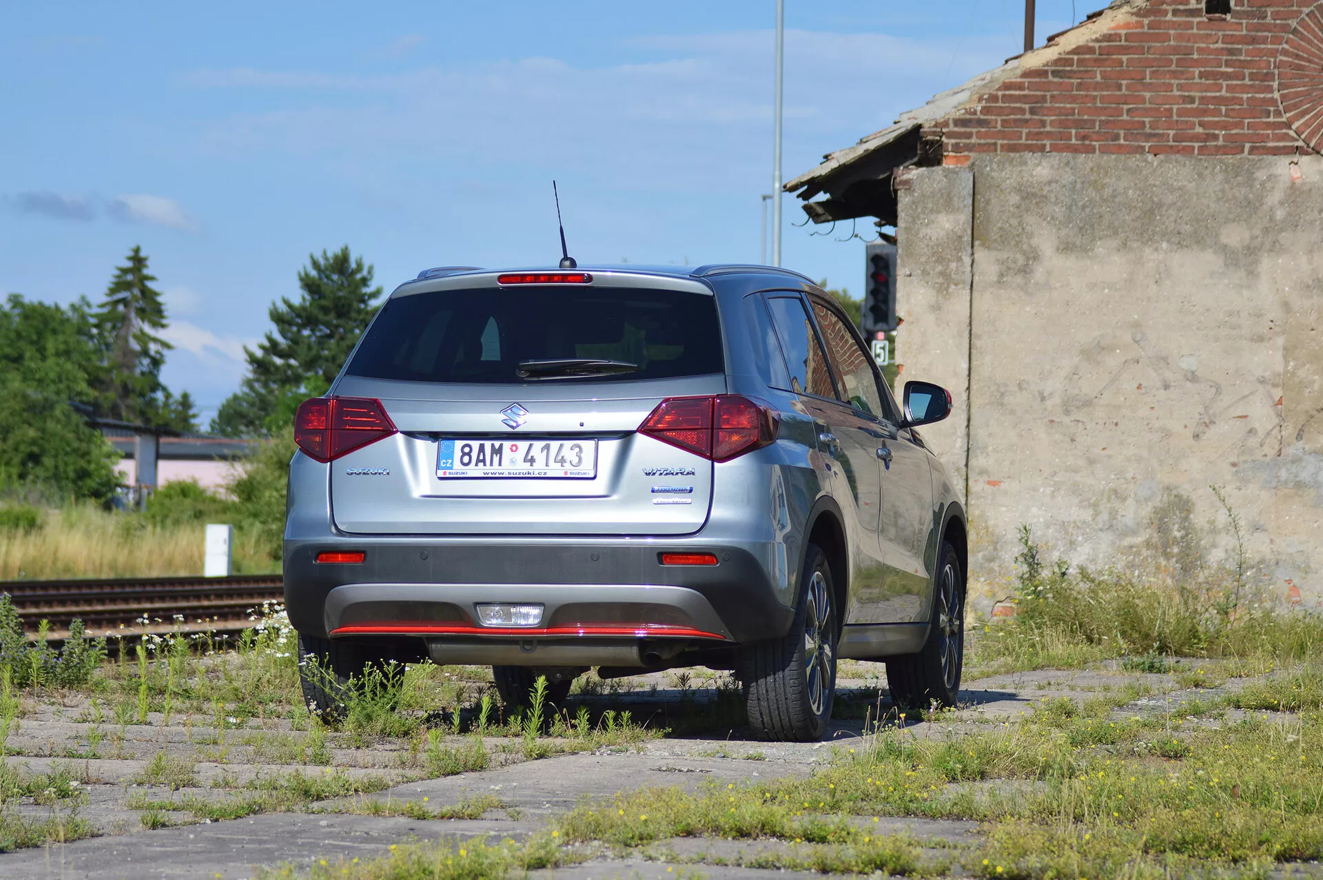 Um SUV compacto Škoda Yeti cinza metálico estacionado em um lote abandonado com manchas de grama e concreto, fotografado de uma vista traseira de três quartos. A imagem mostra o design traseiro distintivo do veículo com luzes traseiras convencionais, limpador de para-brisa traseiro, luz de freio montada no alto e parte de um aerofólio traseiro. O logotipo da Škoda está centrado na tampa traseira, com o nome do modelo 'Yeti' à esquerda e um emblema '4x4' à direita, indicando tração nas quatro rodas. O carro possui rodas de liga leve de cinco raios, parece estar em bom estado e está localizado em uma área industrial com trilhos de trem desativados e uma estrutura dilapidada ao fundo, sob luz solar natural.