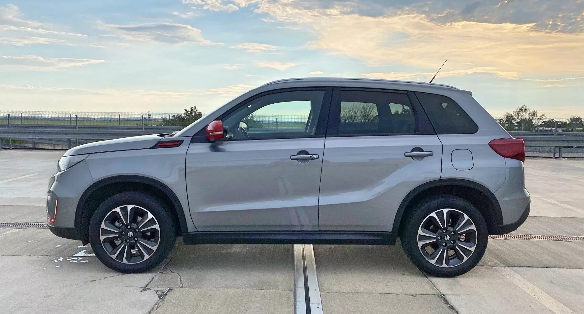 Imagem de perfil lateral de um SUV moderno, Suzuki Vitara 2018, tirada durante o dia em um estacionamento espaçoso. A seção central do veículo é destacada, mostrando o comprimento e os elementos de design lateral, como as formas das portas, soleiras laterais e arcos das rodas. A iluminação natural destaca suas cinco portas, rodas de liga leve de múltiplos raios, barras de teto, maçanetas e elementos decorativos contrastantes, antena e possíveis janelas escurecidas. O SUV está em condição impecável com um brilho refletivo no exterior.