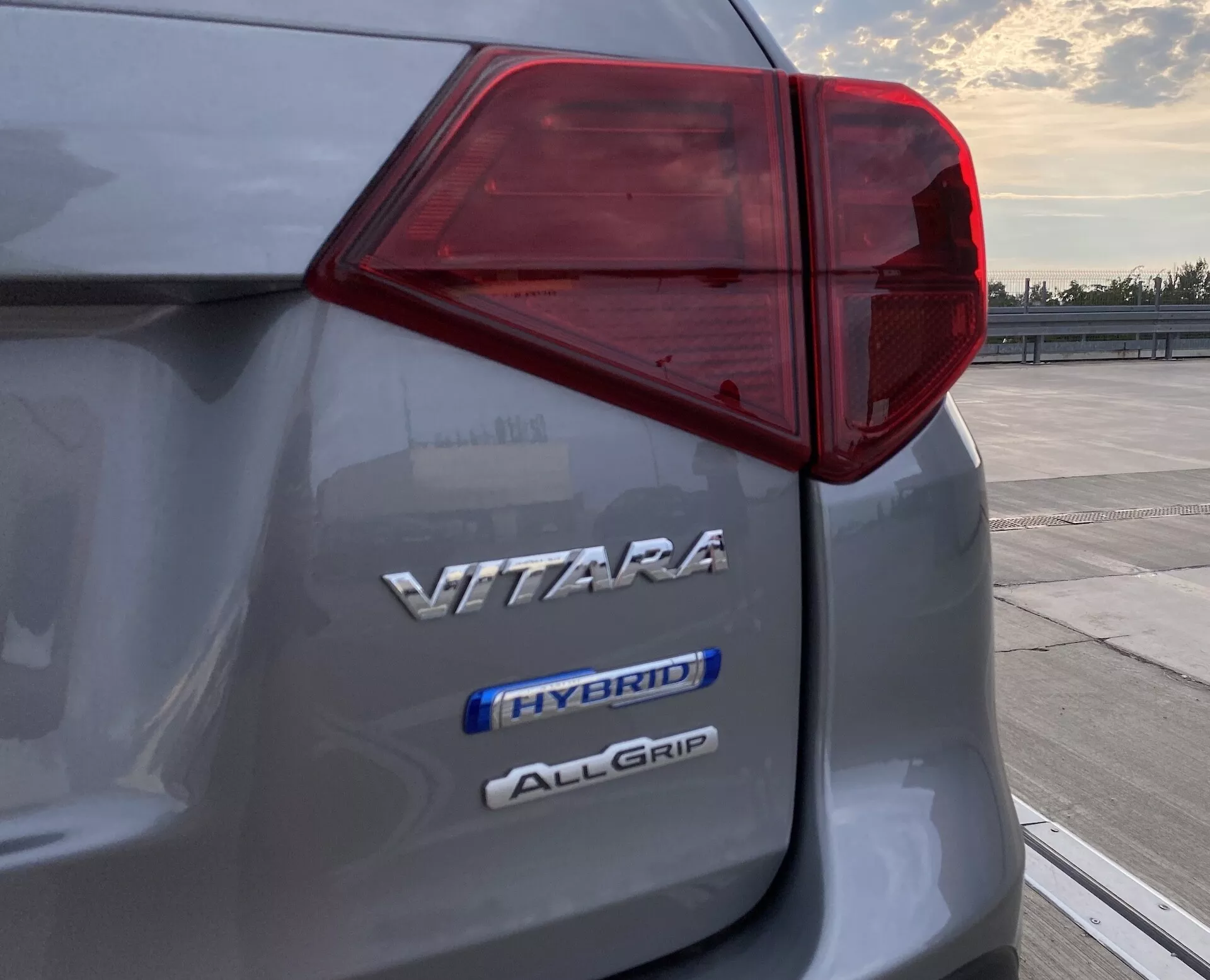 Close-up do aspecto traseiro esquerdo de um Suzuki Vitara 2018 mostrando o design intrincado da luz traseira LED e três emblemas indicando o nome do modelo 'VITARA', tecnologia híbrida 'HYBRID' e o sistema de tração integral 'ALLGRIP', capturado ao amanhecer ou anoitecer em uma área de estacionamento aberta.