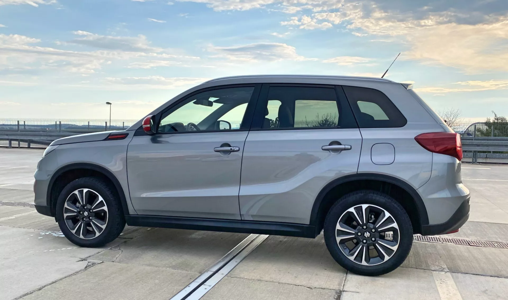 Vista de perfil lateral de um Suzuki Vitara 2018 estacionado no deck superior de uma estrutura de estacionamento sob um céu claro. O SUV apresenta rodas de liga leve de cinco raios, pintura prateada, sinais de volta integrados nos retrovisores laterais, trilhos de teto, maçanetas da porta da cor da carroceria, um degrau lateral sutil e janelas escurecidas com moldura cromada. O design do veículo enfatiza a elegância esportiva e a utilidade, com um corpo quadrado para maximizar o espaço interno. O ambiente exterior e a composição destacam o estilo e a construção do carro, adequado para categorias relacionadas a veículos.