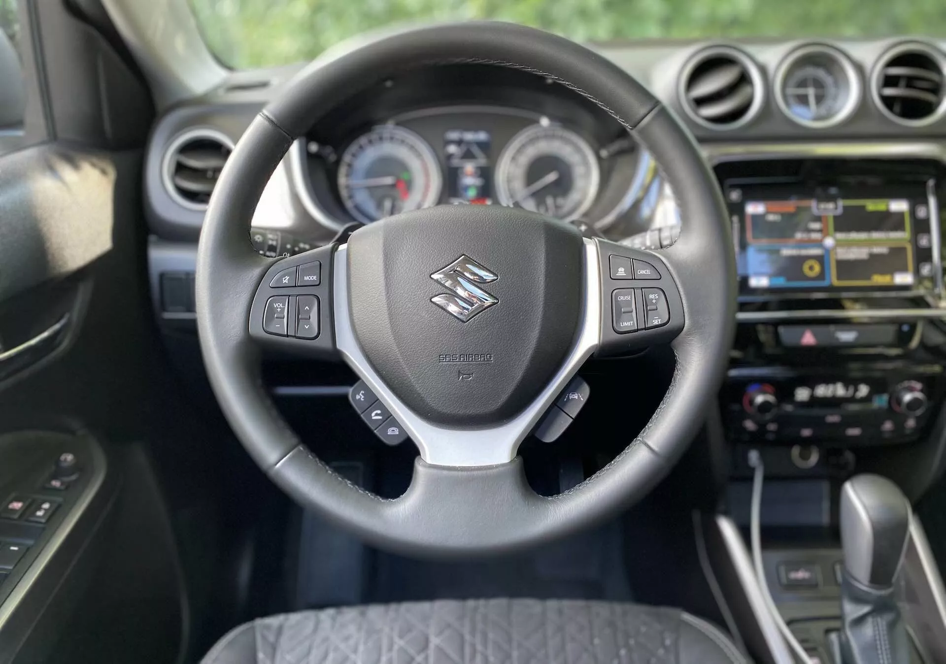 Vista interior de um Suzuki Vitara 2018 a partir da perspectiva do banco do motorista. O foco está no volante, que apresenta o logotipo da Suzuki e controles multifuncionais para áudio, comandos de voz, controle de cruzeiro e gerenciamento da tela de informações do veículo. Atrás do volante, são visíveis mostradores analógicos e uma tela central no painel de instrumentos. A consola central exibe um sistema de infotainment com ecrã táctil com navegação, multimédia e opções de conectividade. Abaixo disso, encontram-se botões táteis e botões para controle climático de zona dupla. O carro está equipado com transmissão automática, indicando um design de cabine moderno e possivelmente premium.