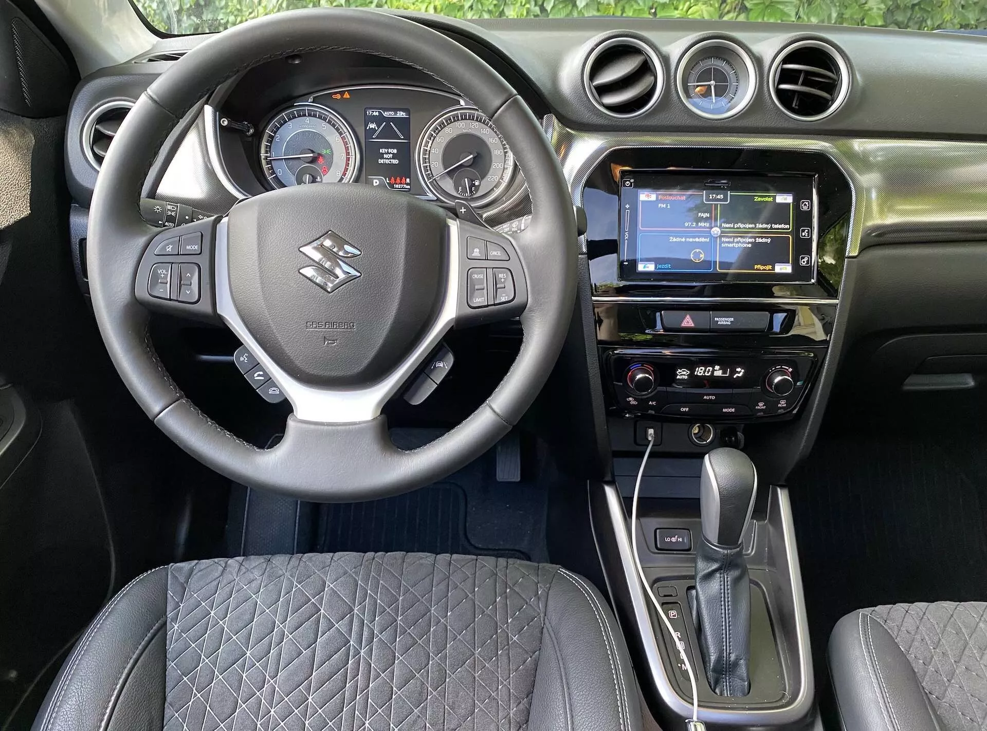 Vista interior de um Suzuki Vitara 2018 a partir do banco do motorista, mostrando o volante central com o logotipo da marca, botões de controle para volume, comando de voz e conectividade telefônica. Atrás dele, um painel de instrumentos digital-analógico com velocímetro, tacómetro e um display digital central. O painel apresenta uma tela de entretenimento multimídia com navegação GPS, rádio e sistema de controle climático. A alavanca de câmbio automático e o freio de estacionamento eletrônico são destacados, junto com um design de assento acolchoado e saídas de ar circulares, capturando a tecnologia automotiva moderna e o design ergonômico.