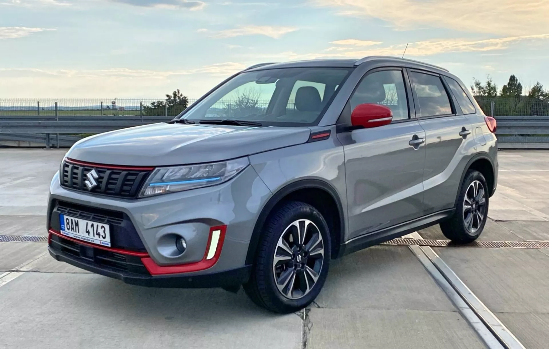A imagem mostra um SUV compacto Suzuki Vitara 2018 estacionado em um ambiente externo em um deck de estacionamento durante o amanhecer ou entardecer. A fotografia captura o carro de um ângulo frontal de três quartos, revelando o lado do motorista e a frente, destacando a configuração dos faróis, o estilo da grade, o para-choques dianteiro, as rodas e as linhas laterais da carroceria. O veículo apresenta uma pintura em duas cores, com a carroceria em prata e o teto e os espelhos laterais em preto, acentuados por detalhes em vermelho no para-choques dianteiro. Os faróis de LED elegantes, um emblema de marca proeminente em uma grade com padrão e rodas de liga leve com múltiplos raios também são visíveis, mostrando o design moderno e esportivo do veículo.