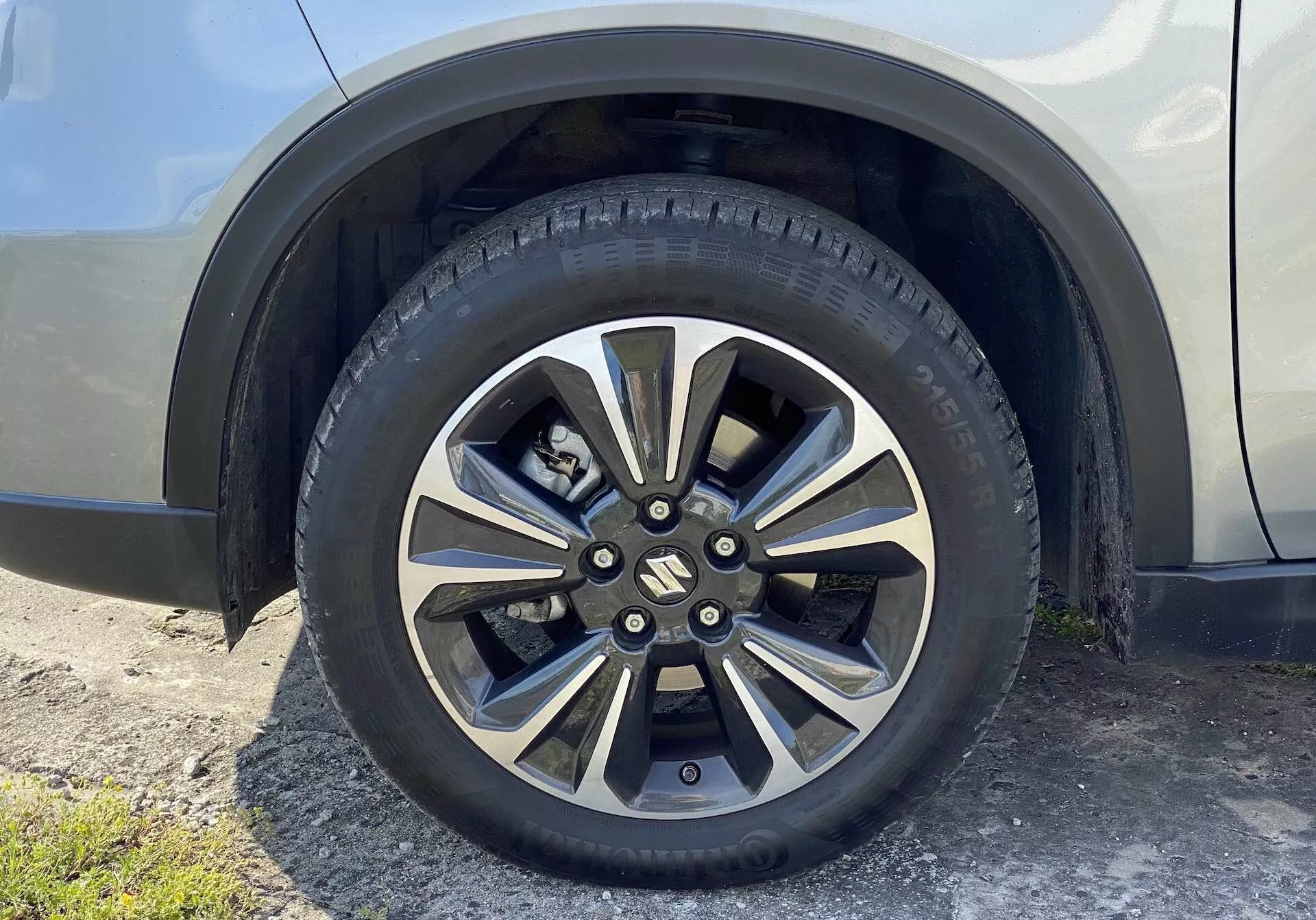 Vista de close da roda do Suzuki Vitara 2018, apresentando um design de liga leve com vários raios metálicos e áreas pintadas mais escuras. A tampa central com marca oculta os parafusos e o cubo do eixo, e o pneu para estrada exibe padrões detalhados de banda de rodagem e marcações na parede lateral. O fundo inclui uma seção do para-lama e do painel lateral do veículo em uma cor neutra e brilhante, vista de um ângulo ligeiramente para cima no nível do solo.