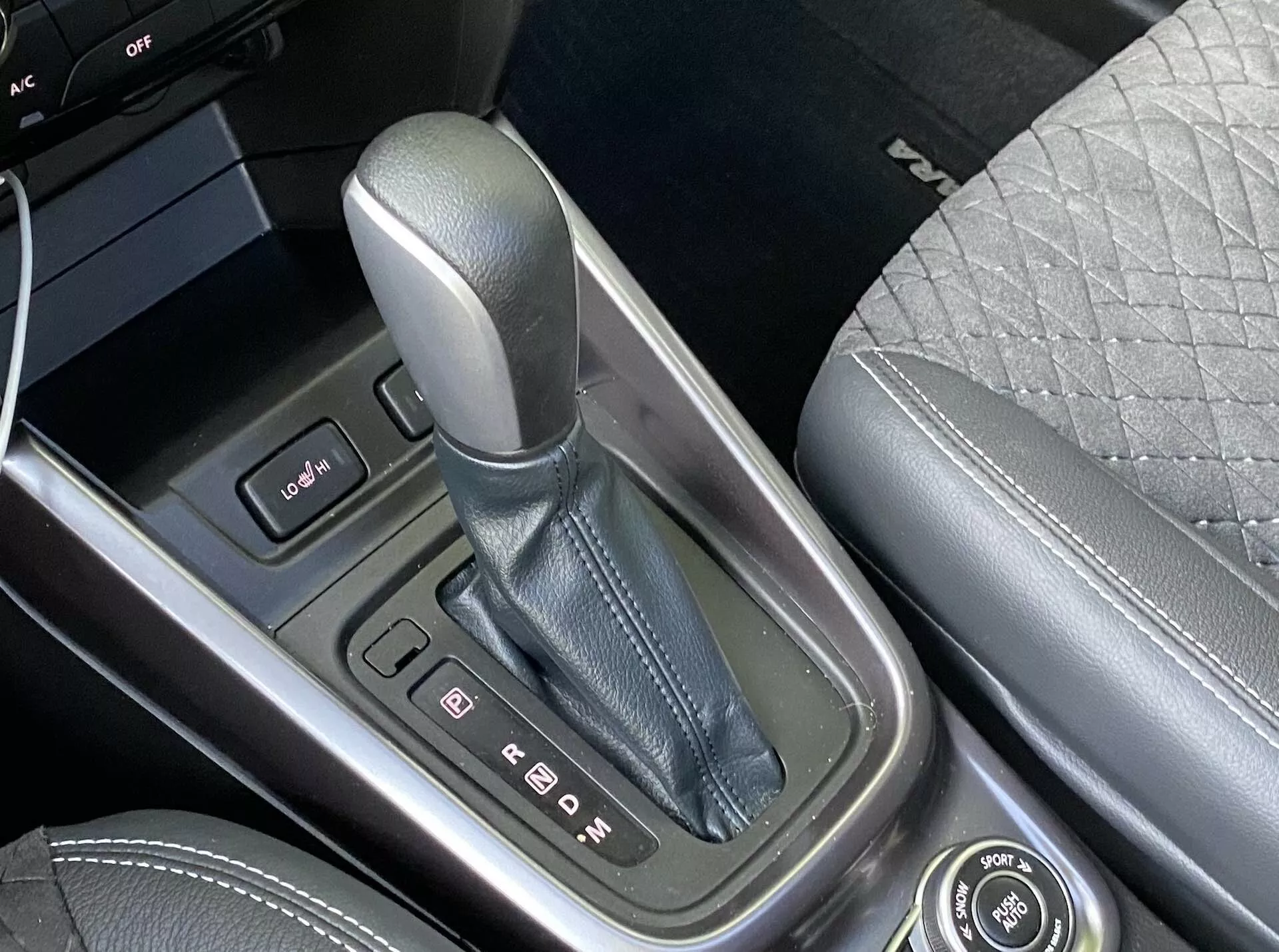 Visão detalhada do console central do Suzuki Vitara 2018, focando na alavanca de câmbio na posição 'P' para estacionamento, circundada por uma moldura com acabamento metálico e indicadores de marchas claros. À esquerda, é visível um painel de controle com dois botões e a borda de uma tela. O console central é feito de materiais de alta qualidade e à direita distingue-se parte do banco do passageiro dianteiro com design acolchoado. Também está presente um botão giratório para funções de infotainment, destacando a tecnologia avançada e os recursos de luxo do veículo.