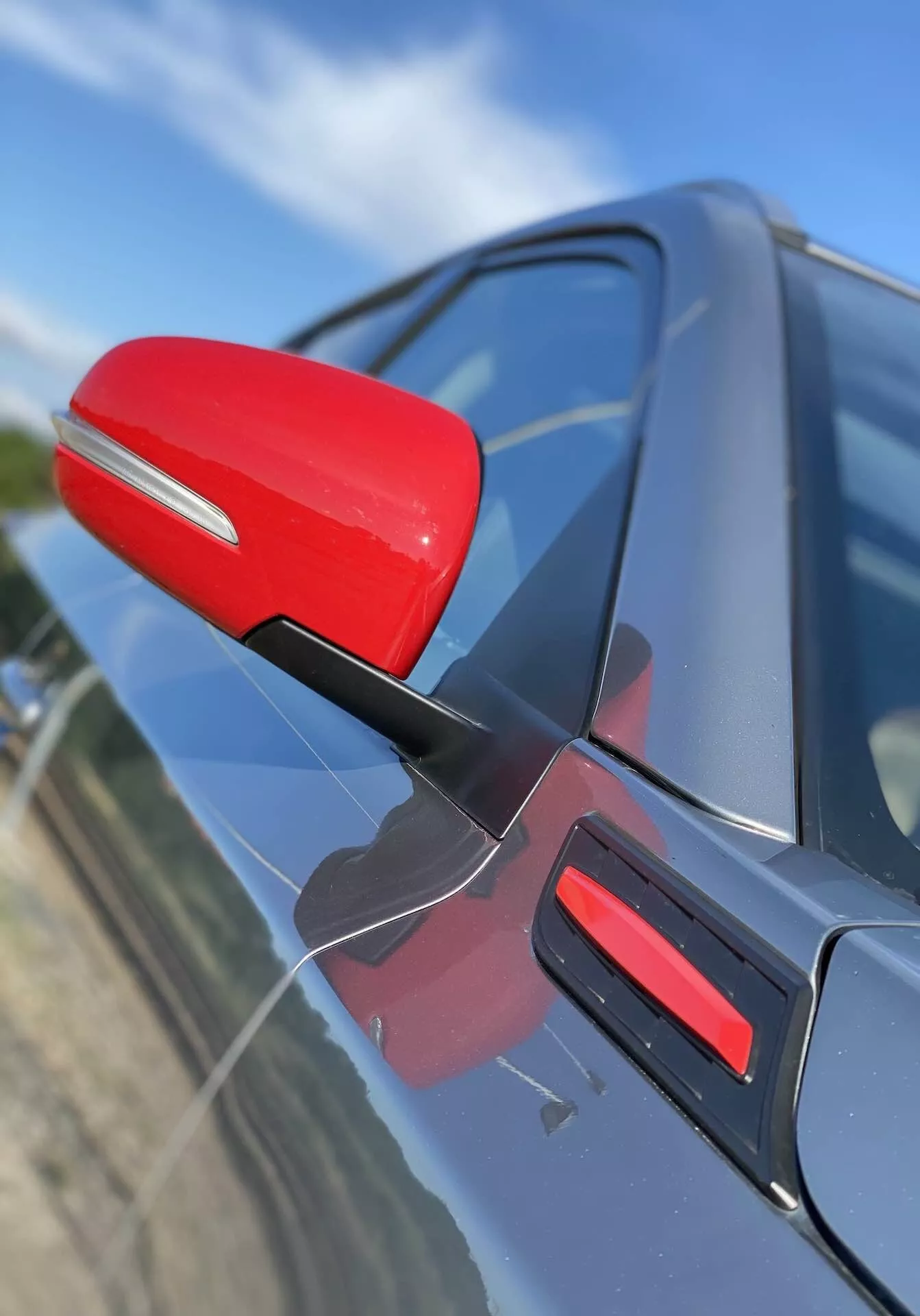 Vista de close-up do espelho lateral do lado do motorista em um Suzuki Vitara 2018, destacado em vermelho contra a carroceria escura do carro. O espelho está fixado com um suporte preto aerodinâmico, sugerindo eficiência aerodinâmica. A foto captura o espelho em um ângulo oblíquo ligeiramente voltado para a frente, com uma perspectiva ligeiramente inclinada para baixo, apresentando um acabamento vermelho correspondente na porta. O fundo está desfocado, enfatizando os detalhes do espelho e do design da porta do carro.