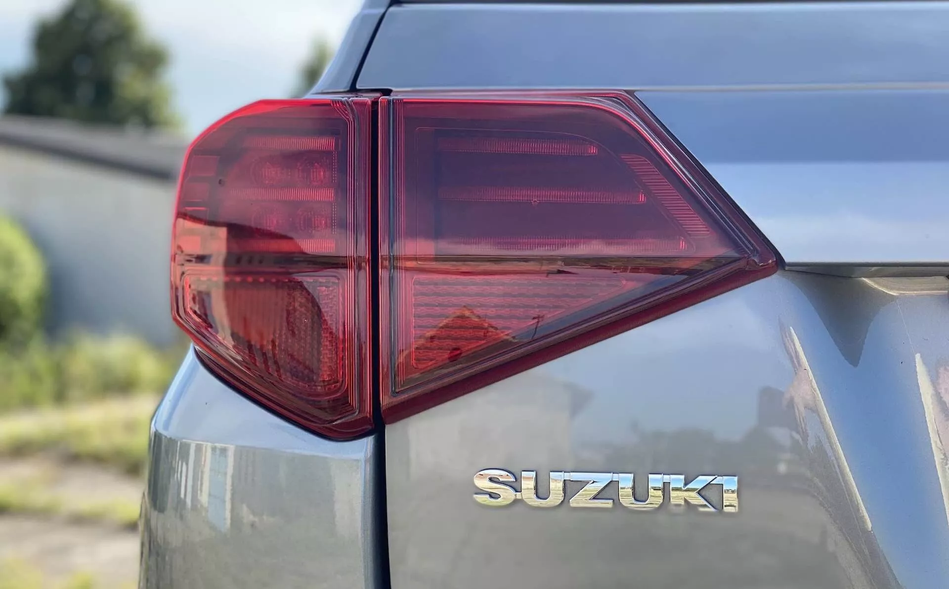 Vista aproximada do lado traseiro esquerdo de um Suzuki Vitara 2018 focalizando na luz traseira. A foto em perspectiva três quartos, tirada de uma altura baixa a média, exibe a luz traseira LED distintamente estilizada com ângulos acentuados e várias câmaras para funções como luz de freio, sinal de seta e possivelmente luz de neblina. A luz predominantemente vermelha, com uma seção transparente, provavelmente para a luz de ré, destaca a tecnologia moderna de iluminação automotiva. A tampa traseira possui uma inscrição cromada 'SUZUKI' centralizada abaixo da janela traseira, enfatizando uma marcação limpa e suave. O acabamento metálico da pintura reflete um pouco de luz, indicando uma superfície do veículo bem mantida ou mais nova.
