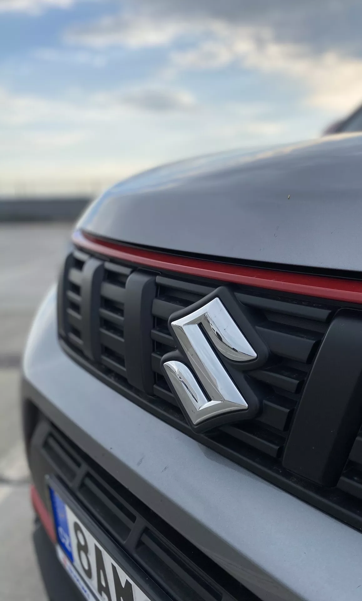 Close-up, visão em ângulo baixo da seção frontal de um Suzuki Vitara 2018. A imagem destaca o emblema do fabricante centrado na grade, que possui lâminas horizontais e um acabamento vermelho ao longo da borda superior, sugerindo um pacote de design mais esportivo. O capô e as seções do carro estão desfocados, destacando um acabamento branco ou de cor clara com um vinco perceptível. O fundo também está desfocado, sugerindo um ambiente externo, possivelmente em um estacionamento, durante a manhã cedo ou tarde da tarde.