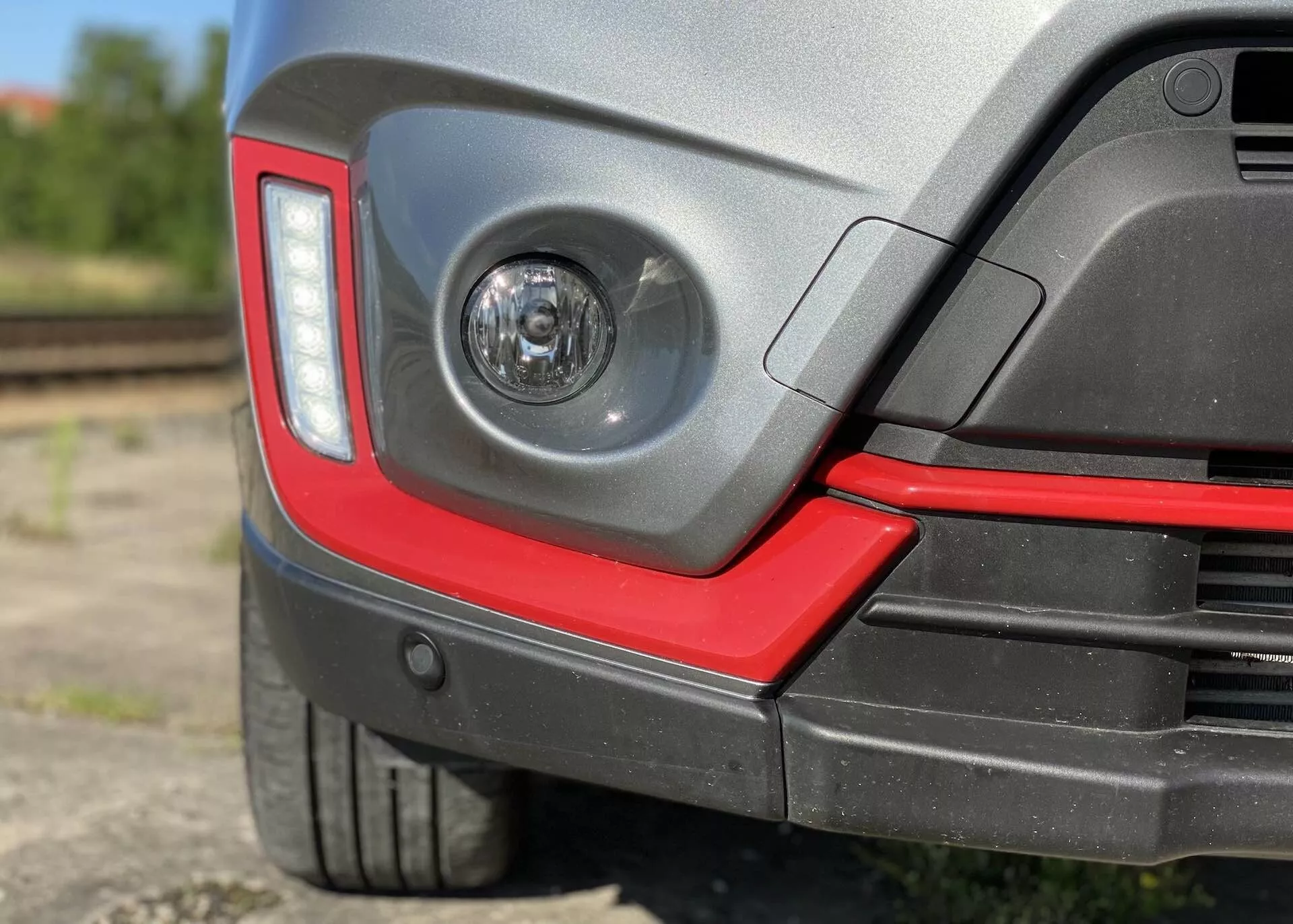 Imagem em close-up da seção traseira de um Suzuki Vitara 2018, destacando um farol de neblina de lente clara no para-choque traseiro, design moderno angular das lanternas traseiras, para-choque traseiro de duas cores com um detalhe vermelho esportivo, possíveis sensores de estacionamento, pneu de perfil baixo com arco de roda de plástico protetor, acabamento de pintura brilhante e fundo desfocado semi-industrial com trilhos ferroviários.